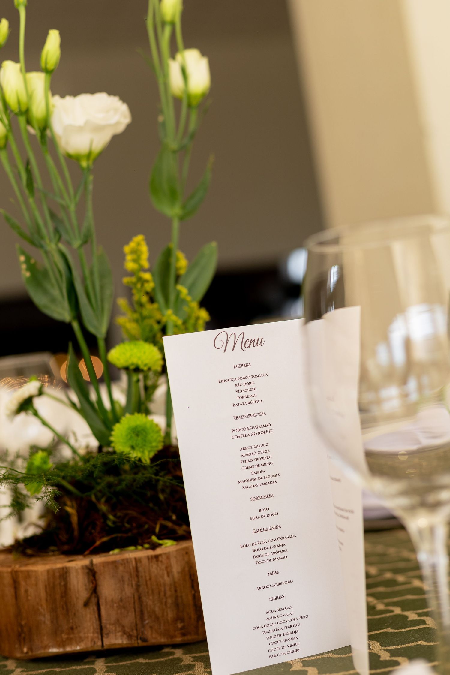Elegant wedding menu card displayed on a table with white floral centerpiece in a wooden base and wine glass.