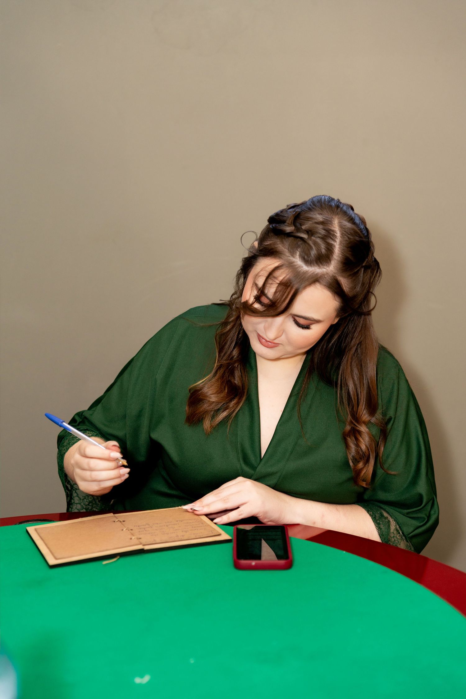 Young woman in green dress writing in notebook at green table with smartphone nearby.