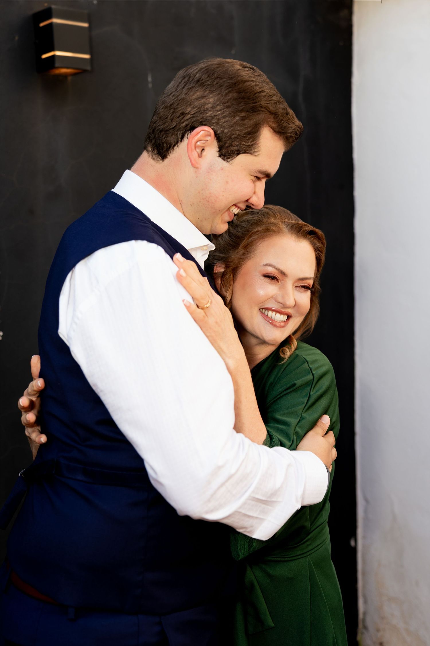 Happy couple embracing outdoors, woman in green dress laughing joyfully, man in white shirt and navy vest holding her close.