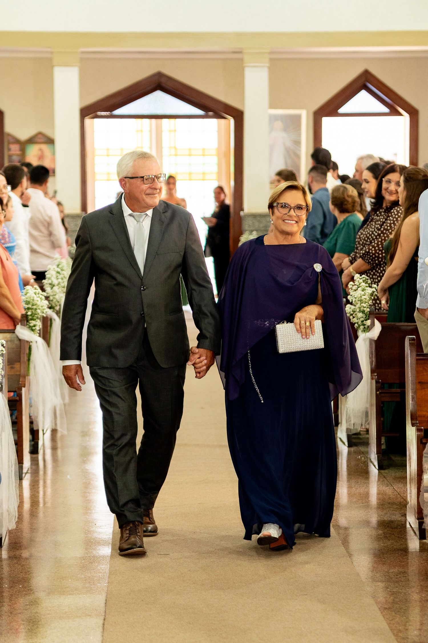 Elegant couple walking hand in hand down church aisle at wedding ceremony, woman in purple dress, man in dark suit.