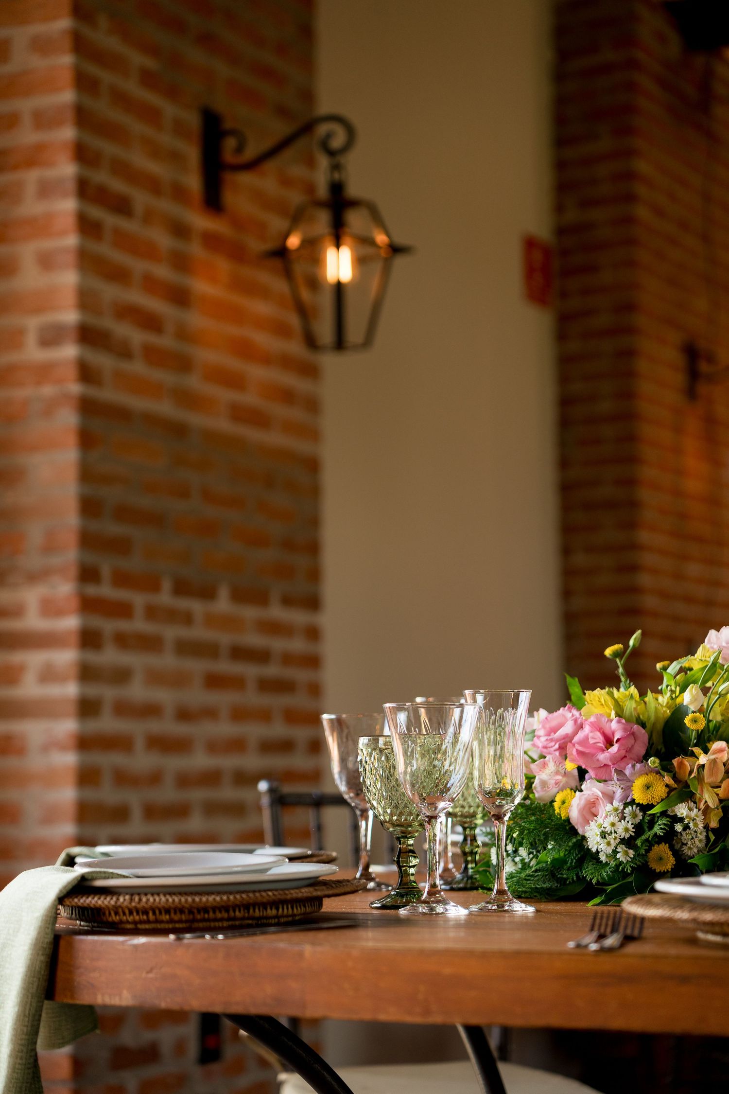 Elegant dining table with crystal glasses, floral centerpiece, and brick wall with vintage lantern in a cozy restaurant setting.