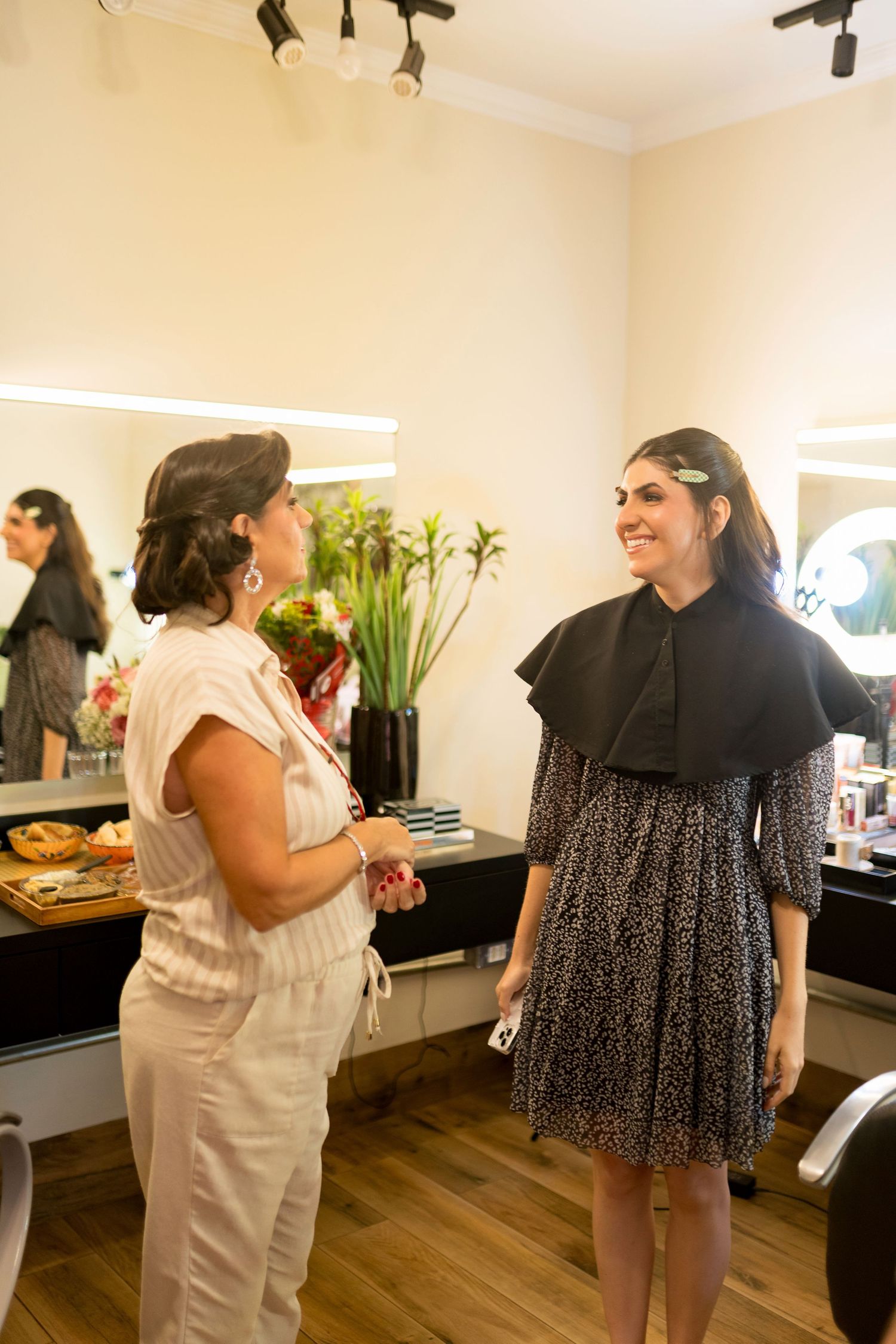 Two women chatting and smiling in a beauty salon, one in white jumpsuit, other in black floral dress with hair accessories.