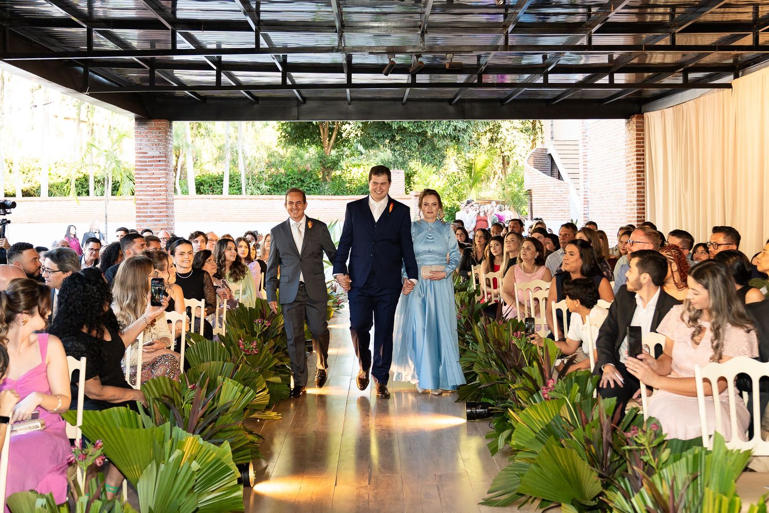 Bride in blue gown walks down wedding aisle with two men at elegant indoor ceremony surrounded by guests.