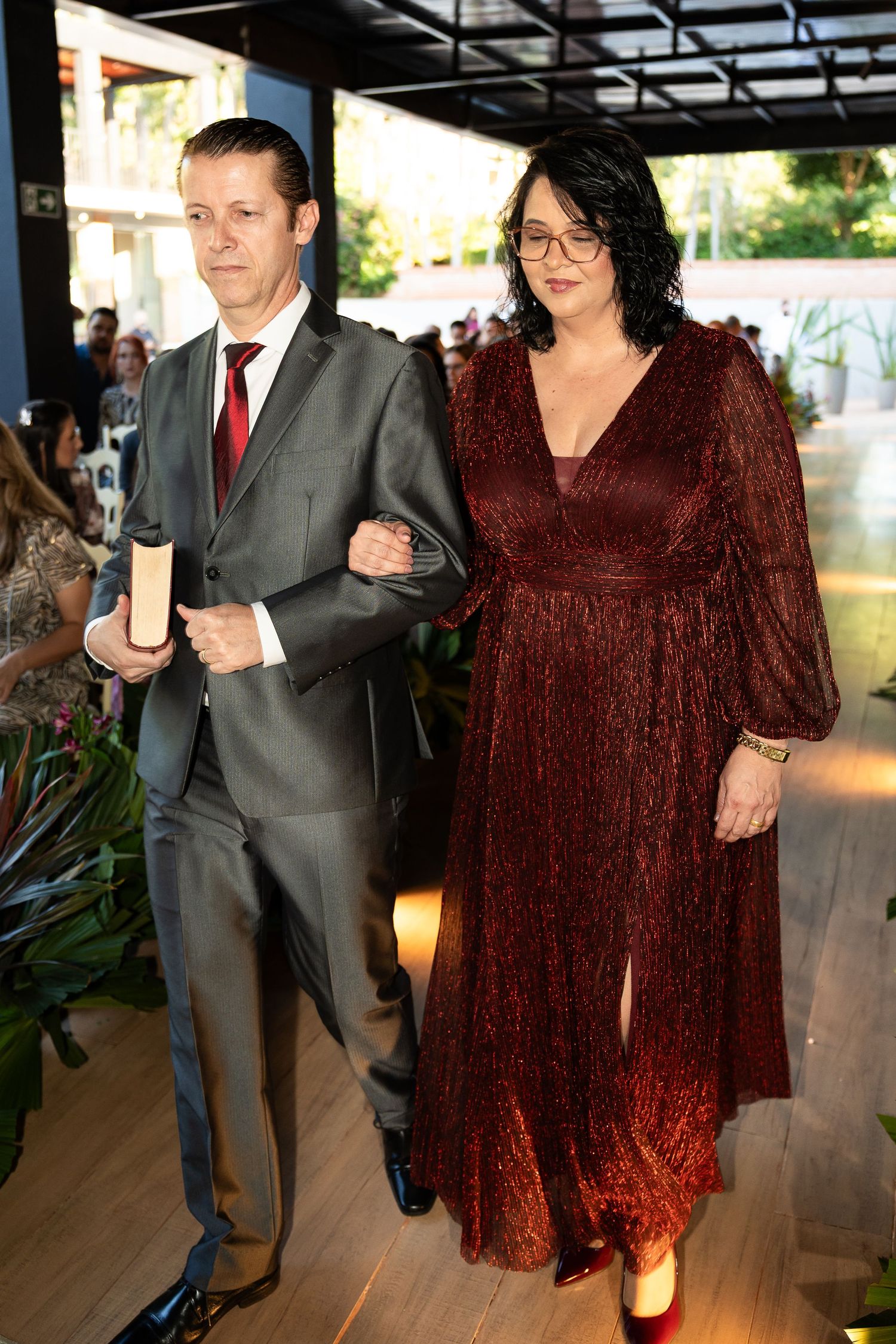 Elegant couple in formal attire, man in gray suit with red tie, woman in burgundy sparkly gown, walking at event.