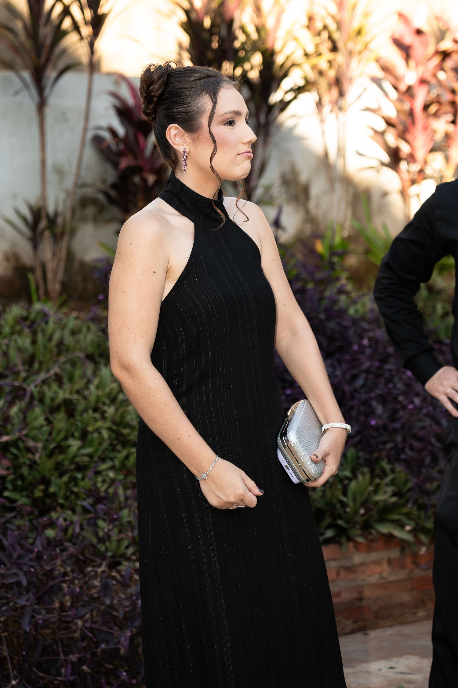 Woman in elegant black halter neck gown holding silver clutch purse at formal outdoor event.