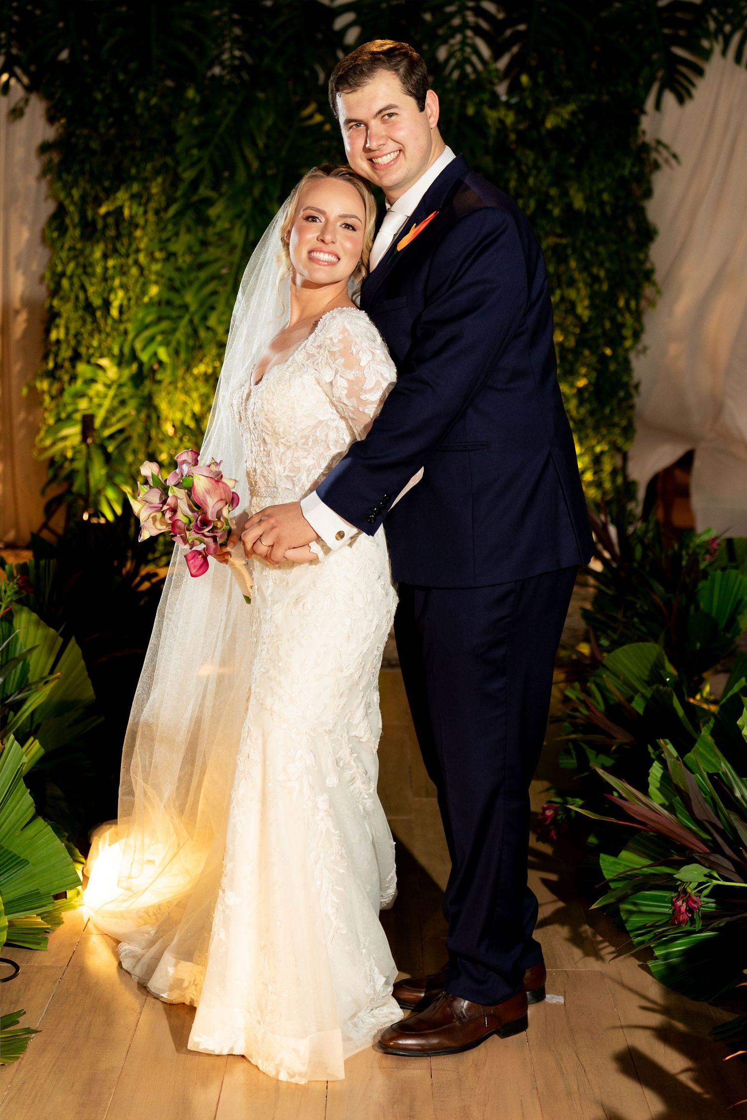 Bride in lace gown with veil holds bouquet beside groom in navy suit, posing before lush greenery backdrop.