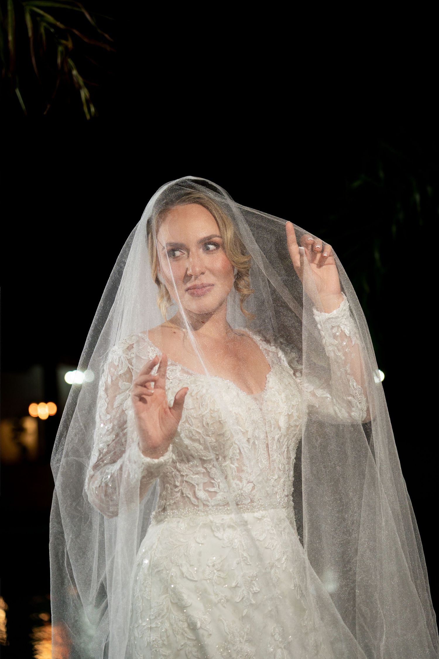 Bride in lace wedding dress lifting sheer veil with both hands, posing outdoors at night.