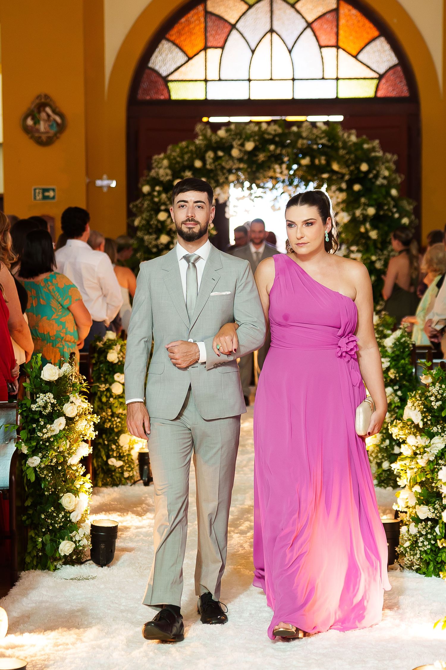 Elegantly dressed couple walks down a floral-adorned church aisle, man in gray suit and woman in purple gown.