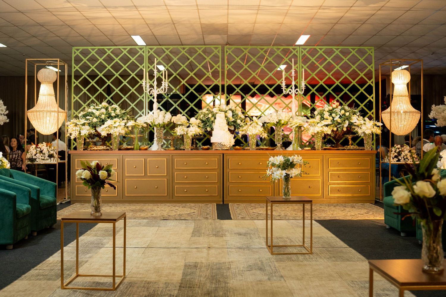 Elegant wedding reception venue with gold lattice backdrop, white floral arrangements, chandelier, and green velvet seating.