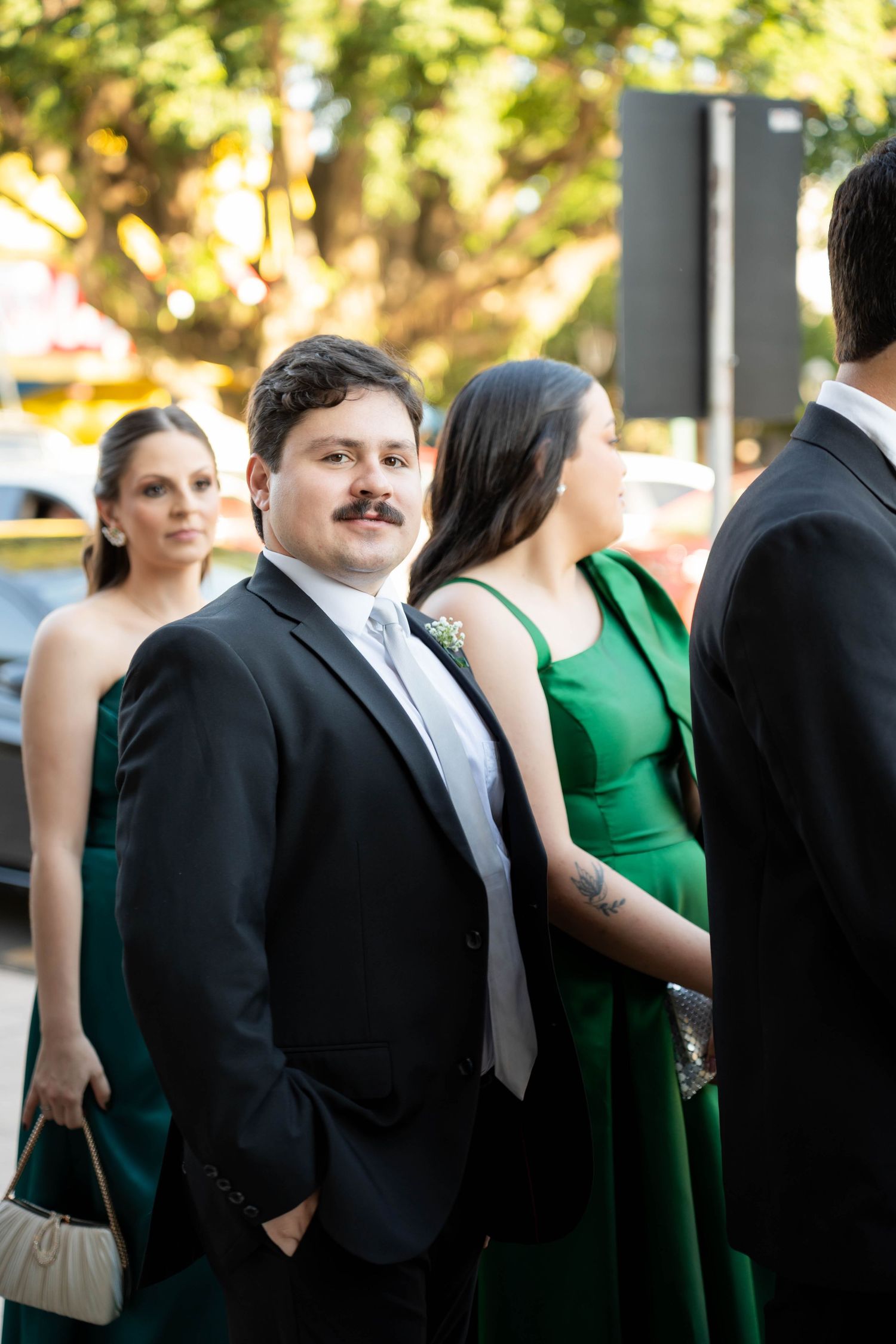 Man in black suit with mustache at formal event, surrounded by women in elegant green dresses outdoors.