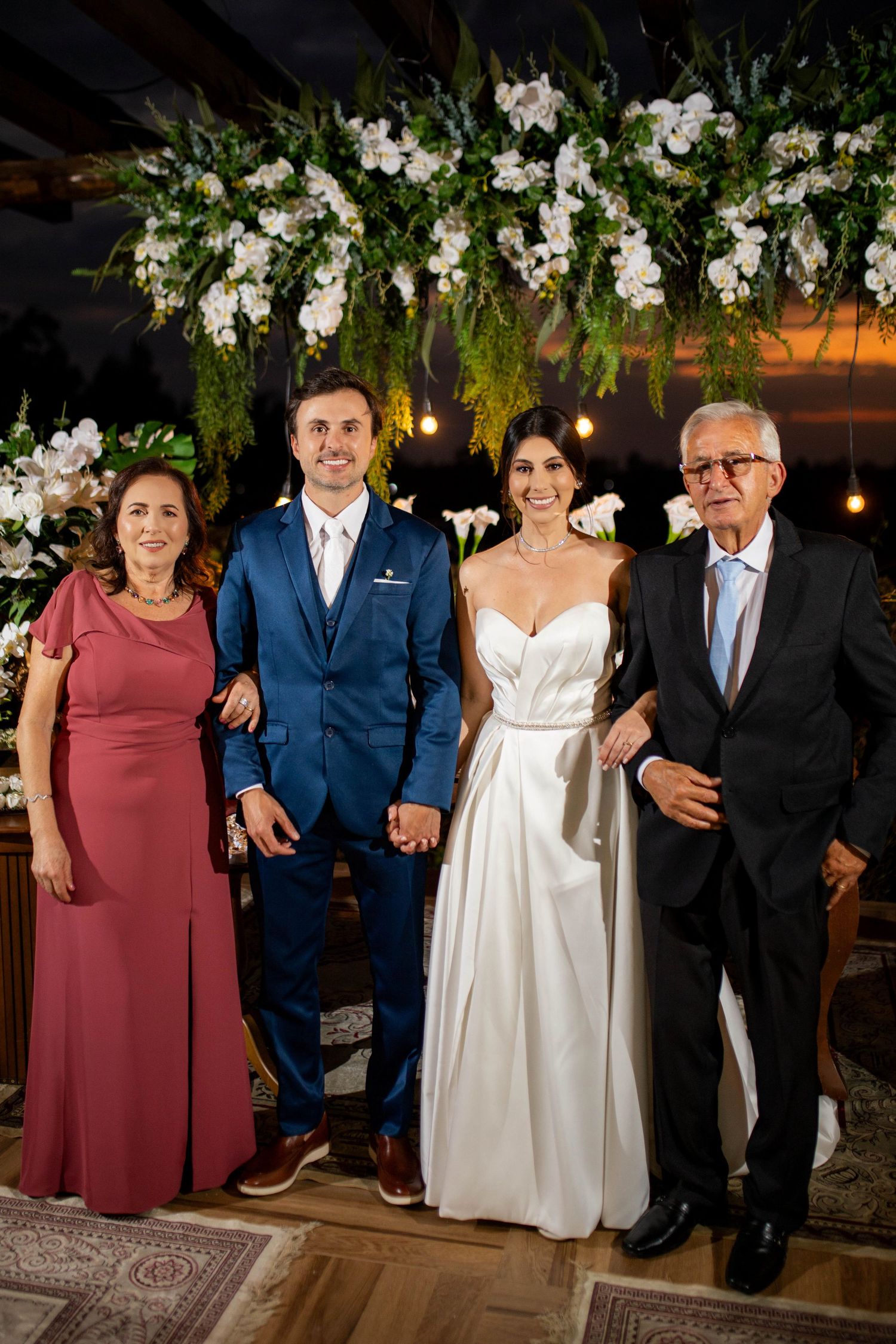 Wedding couple poses with family under floral arch with white orchids and greenery at elegant reception venue.