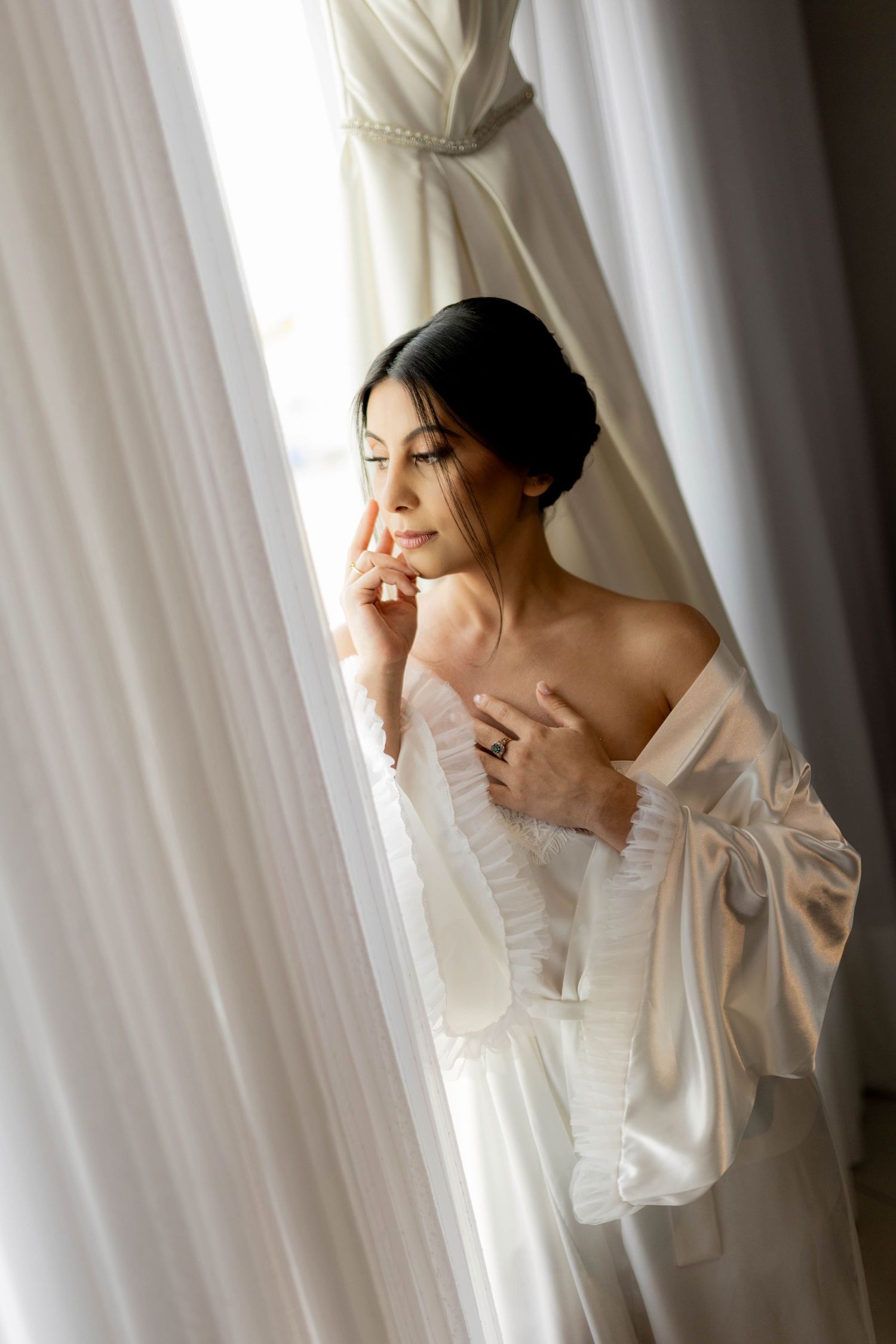 Bride in white satin robe gazing out window with wedding dress hanging in background.