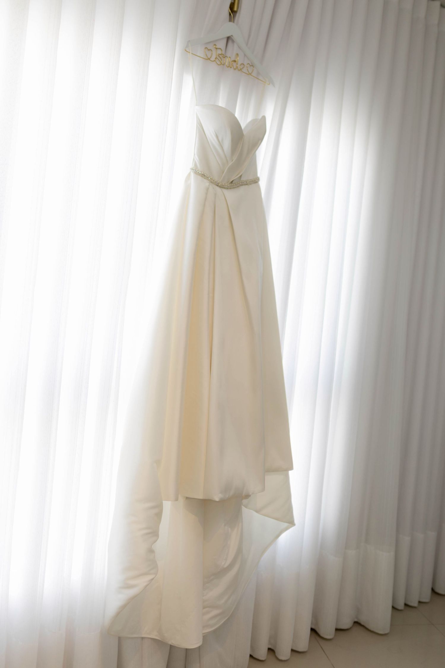 Elegant strapless ivory wedding gown with draped bodice and crystal belt hanging on a hanger by sheer white curtains.