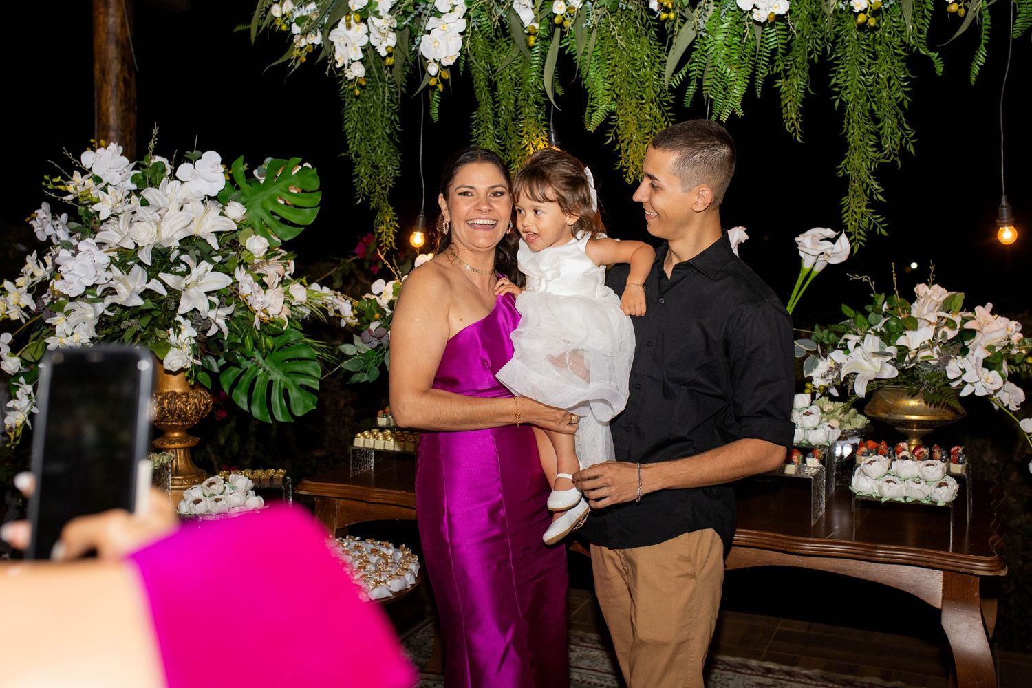A couple in elegant attire holds a young child at a floral-decorated event with white orchids and tropical greenery.