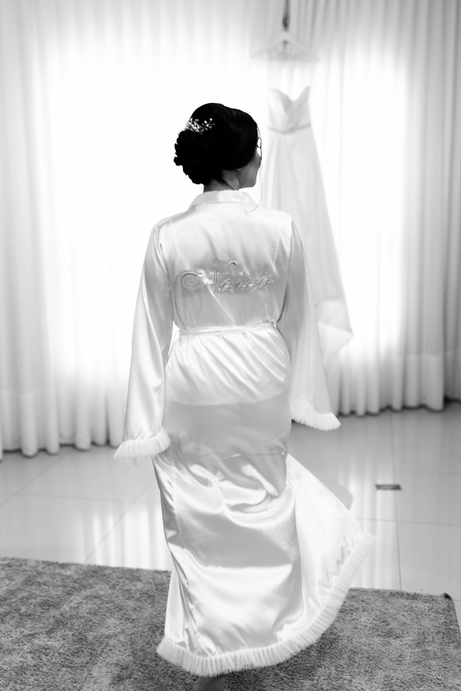 Bride in white satin robe with Bride embroidery stands before window in black and white wedding preparation photo.