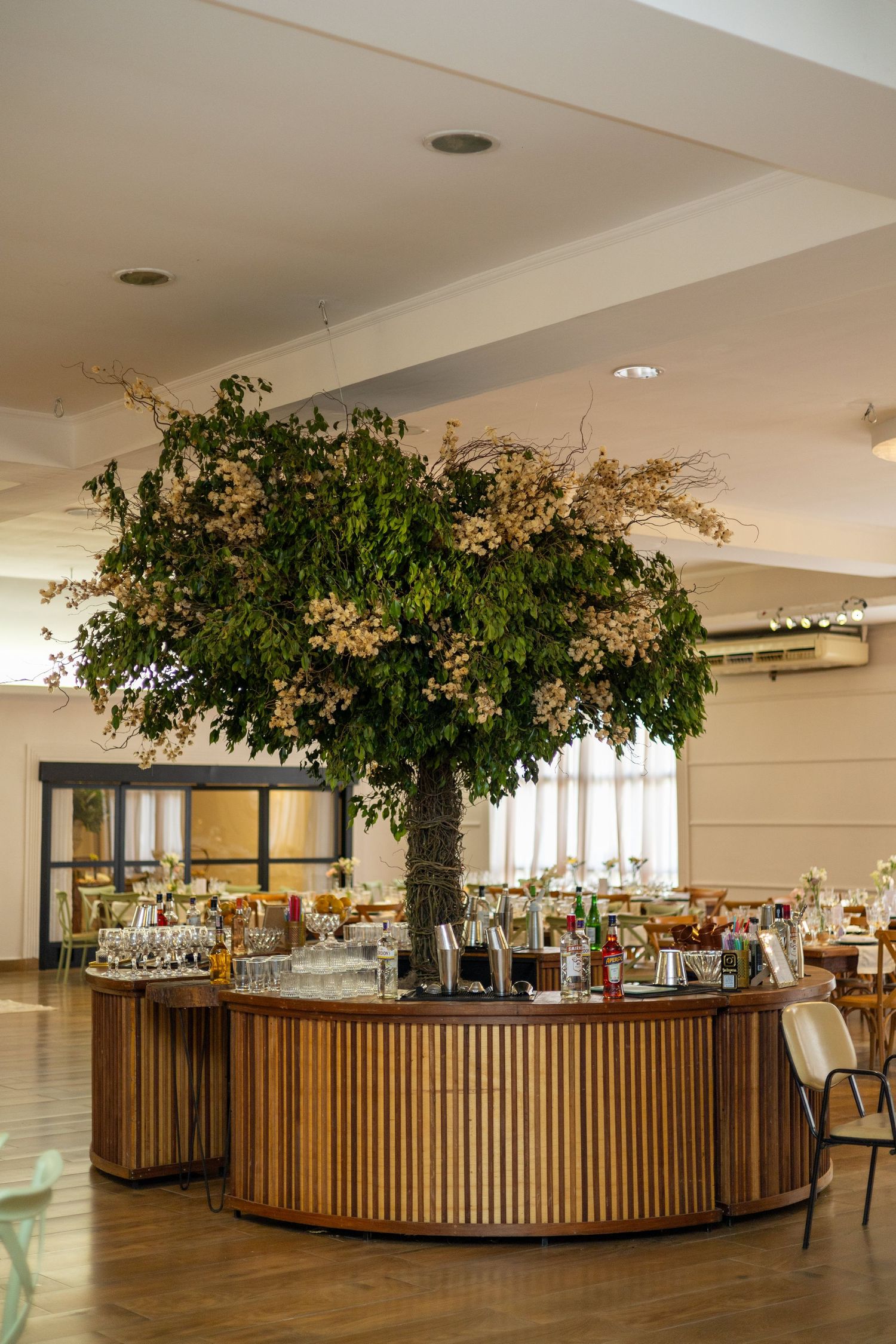 Stunning circular wooden bar with a large lush green tree centerpiece in a bright modern event venue.
