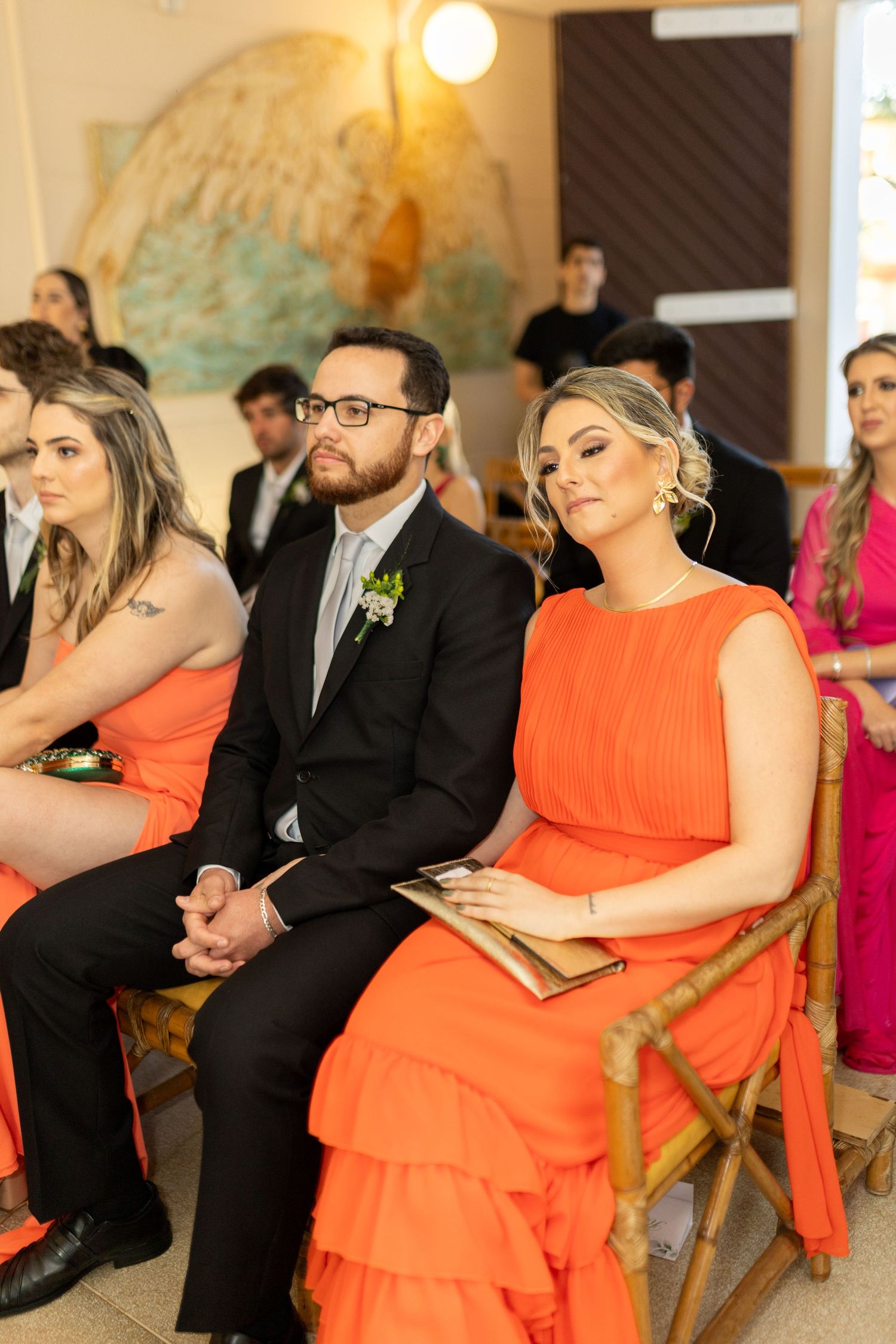 Guests in orange dresses and black suits seated at a wedding ceremony.