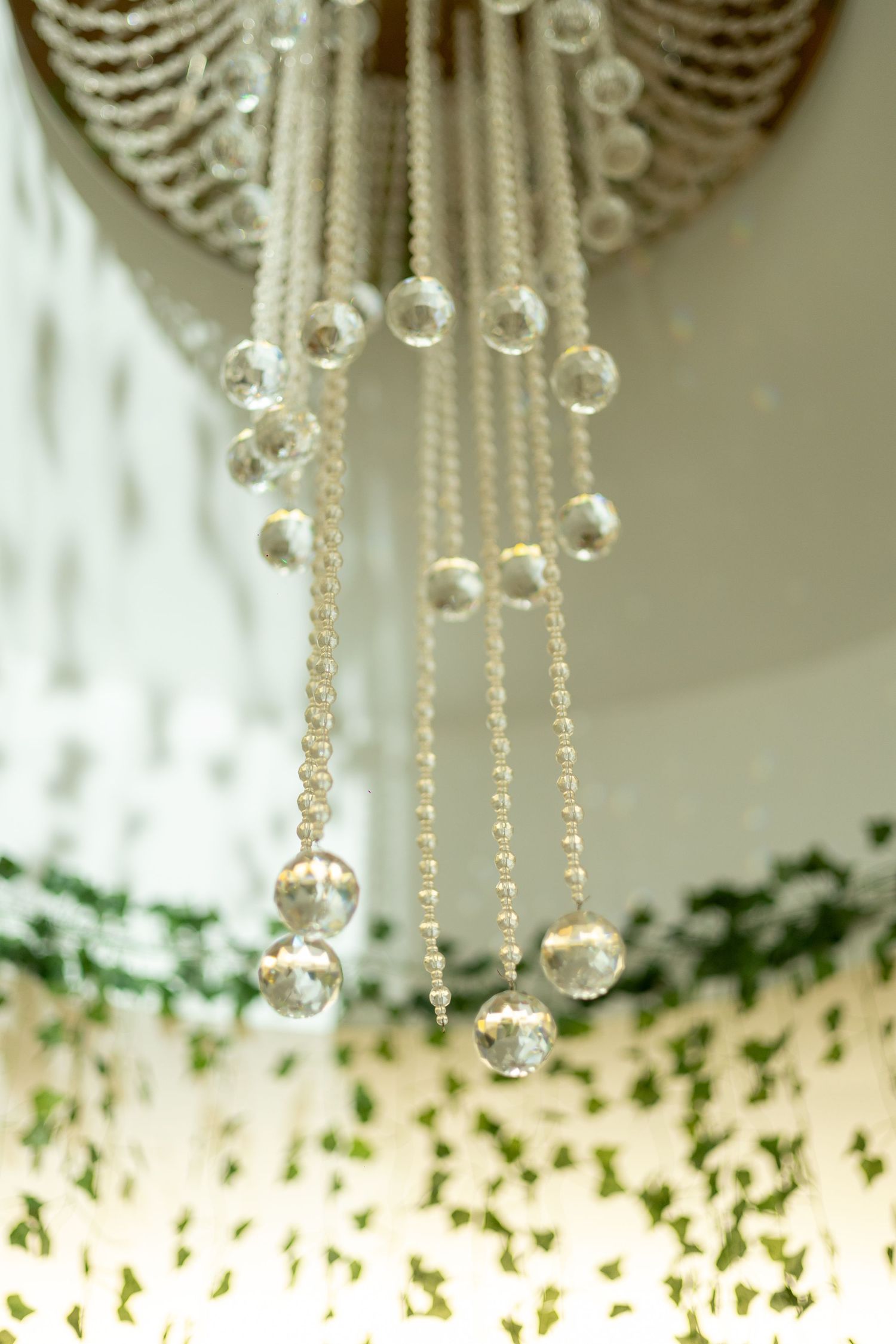 Elegant crystal chandelier with cascading beaded strands and glass ball pendants hanging above lush green foliage.