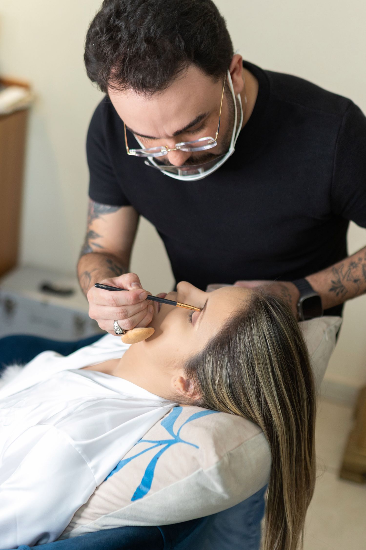 Tattoo artist with glasses applying precise facial makeup or tattoo design on a woman lying in a reclining chair.