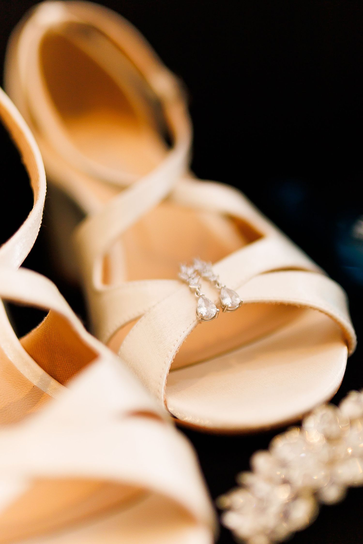 Elegant ivory bridal heels with delicate crystal drop earrings resting on the strap, set against a dark background.
