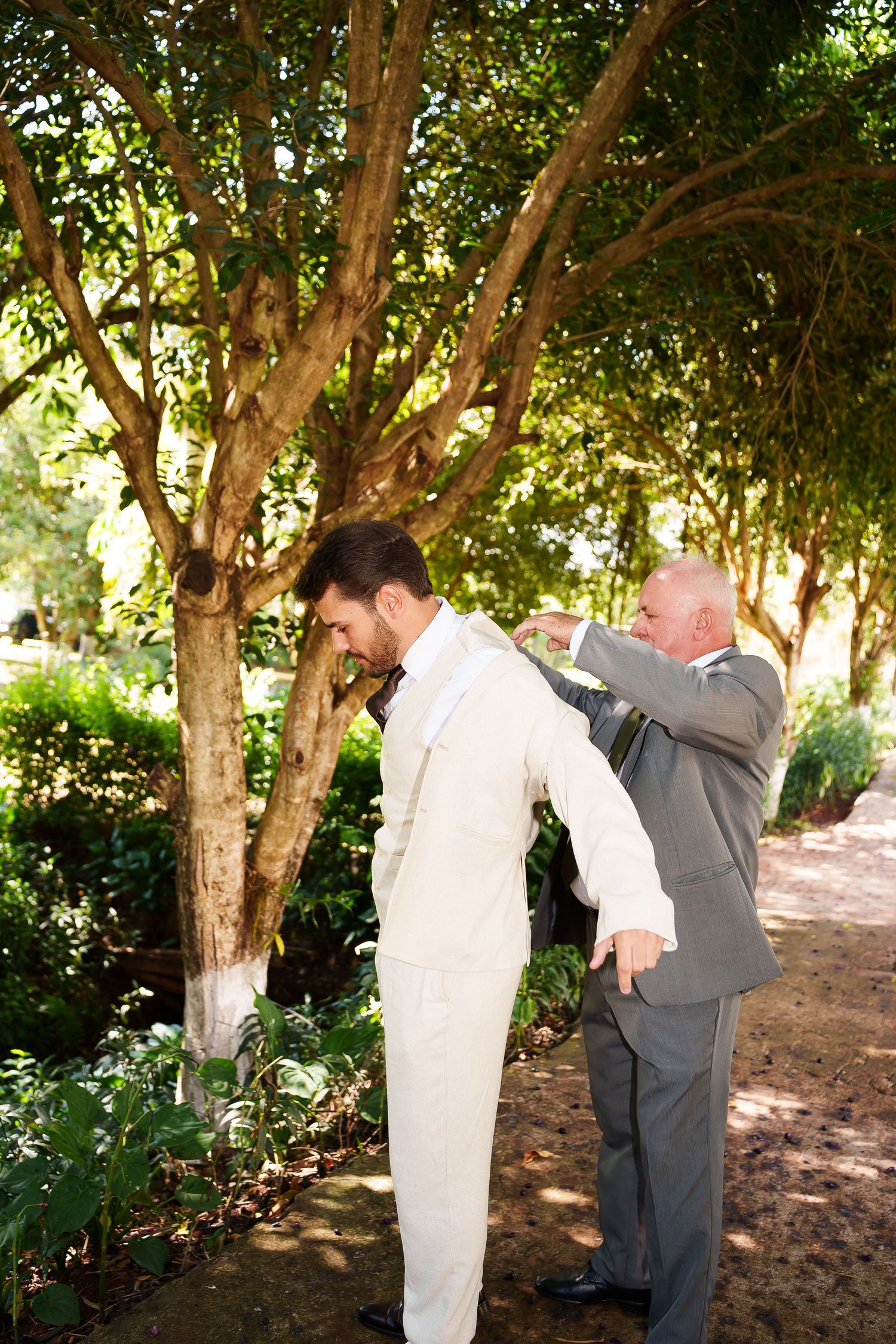 Groom in white suit gets jacket adjusted by older man in gray suit outdoors among lush green trees.