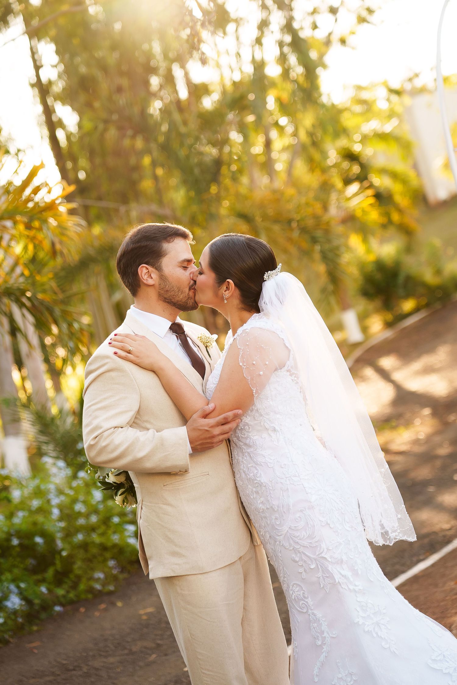 A bride and groom share a romantic kiss outdoors, with the bride in a lace gown and veil and groom in a beige suit.