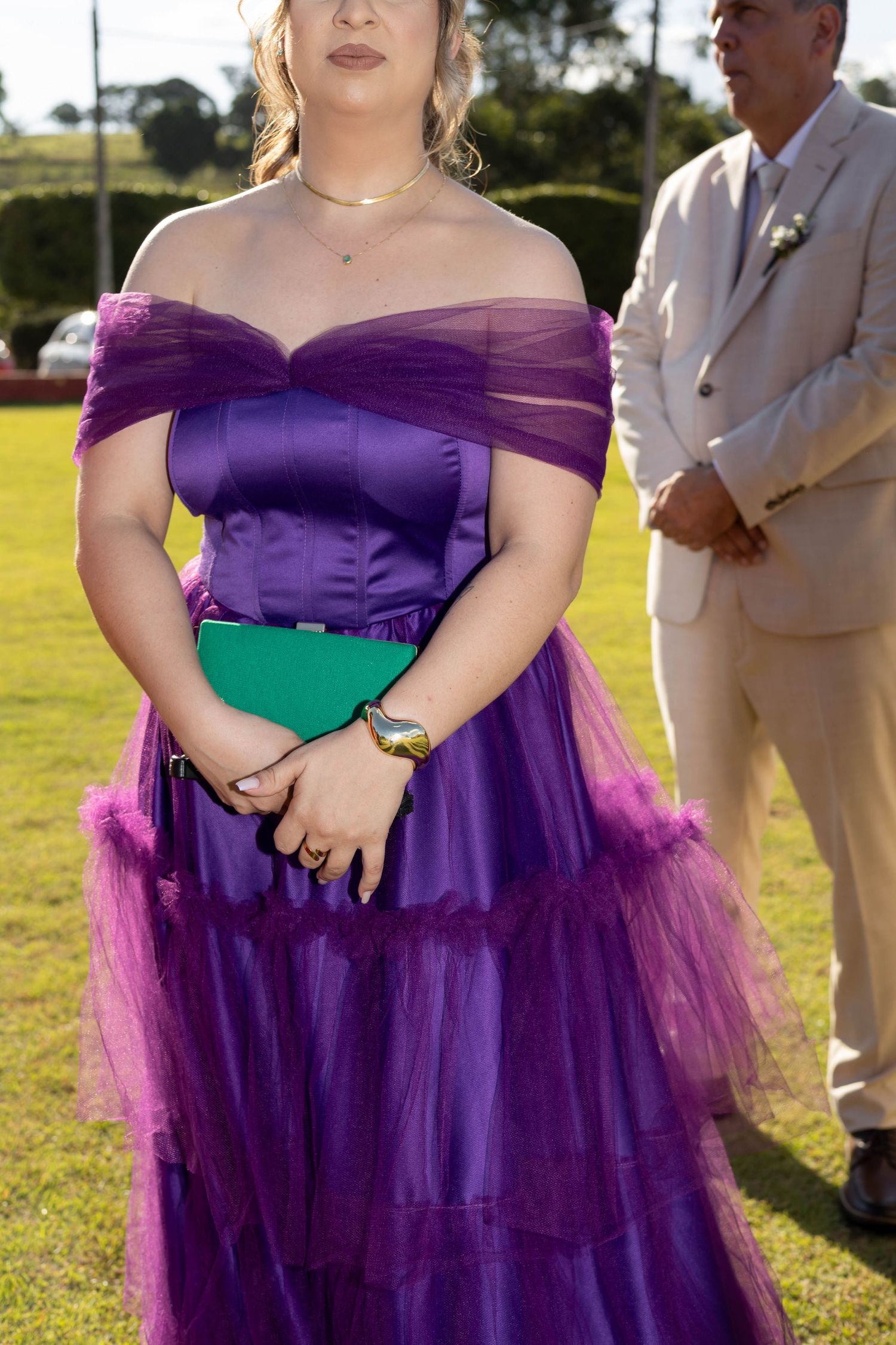 Woman in elegant purple off-shoulder tulle gown holding green clutch at outdoor formal event.