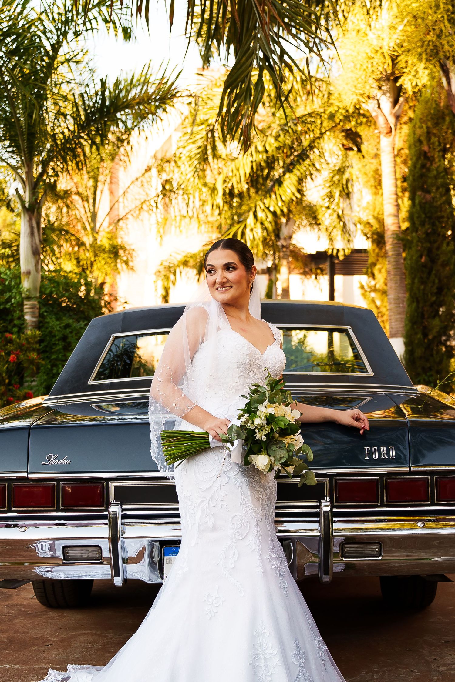 Bride in lace mermaid gown holding bouquet poses beside vintage Ford LTD car surrounded by palm trees.
