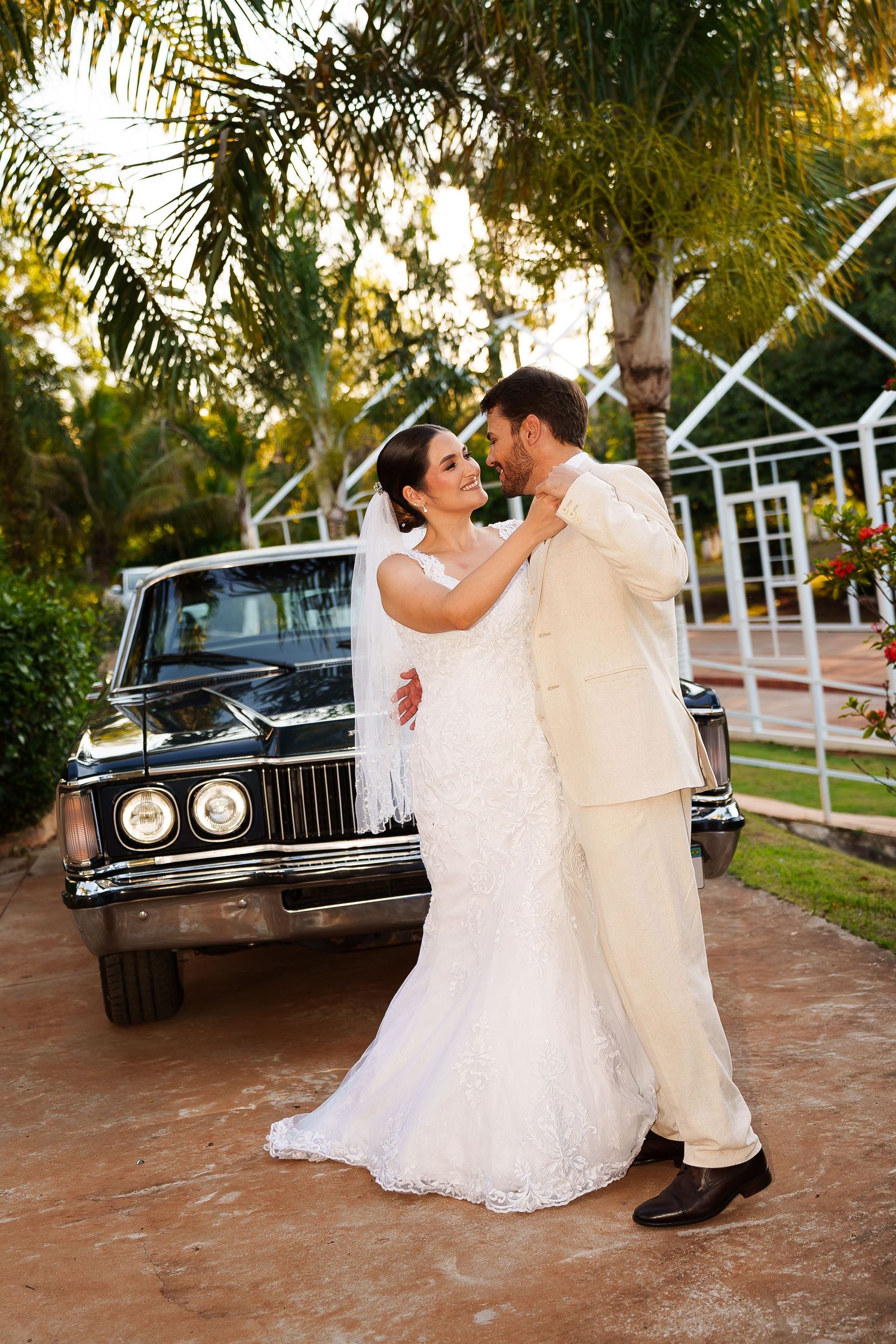 Bride and groom share a romantic moment beside a vintage black car surrounded by tropical palm trees at their outdoor wedding.
