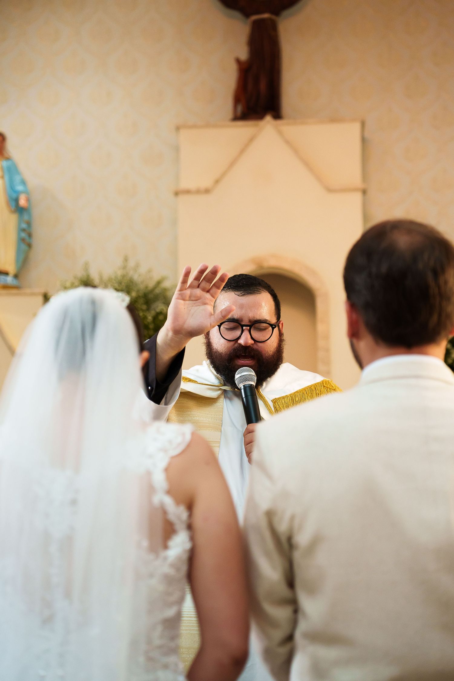 Bearded priest in vestments raises hand in blessing during a Catholic wedding ceremony at the altar.