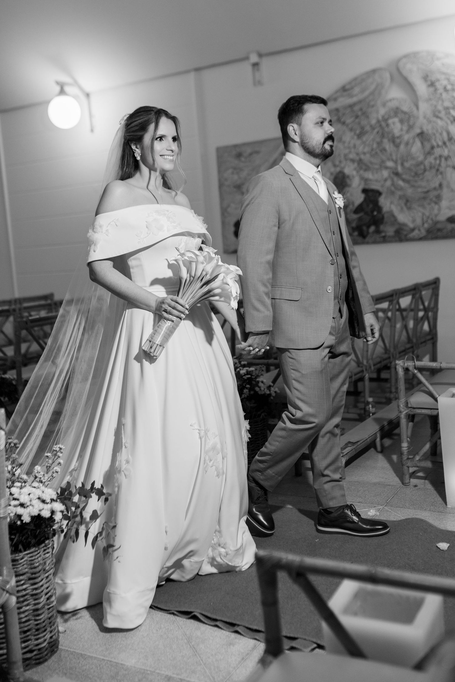 Black and white photo of a bride in off-shoulder gown holding bouquet walking with groom in suit at wedding ceremony.