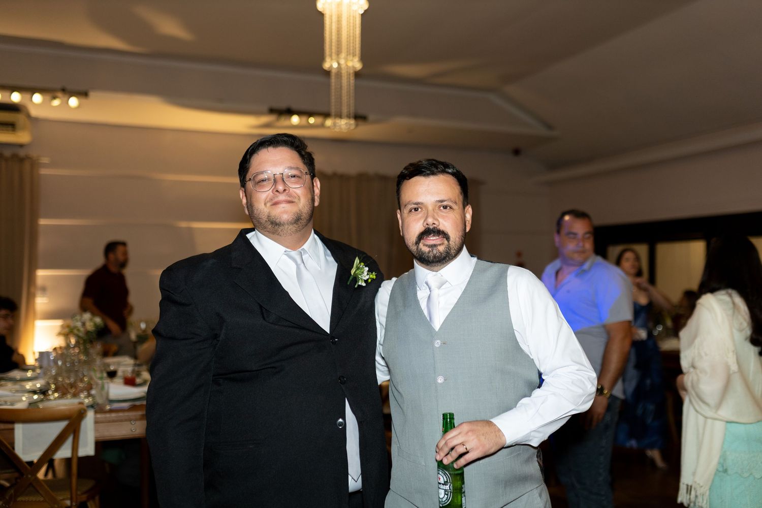 Two men posing together at a wedding reception, one in a black suit and one in a gray vest holding a green bottle.