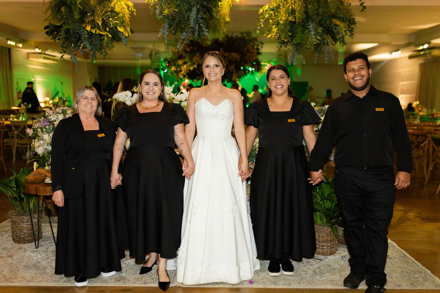 Bride in white gown poses with four wedding staff in black uniforms in an elegantly decorated venue.
