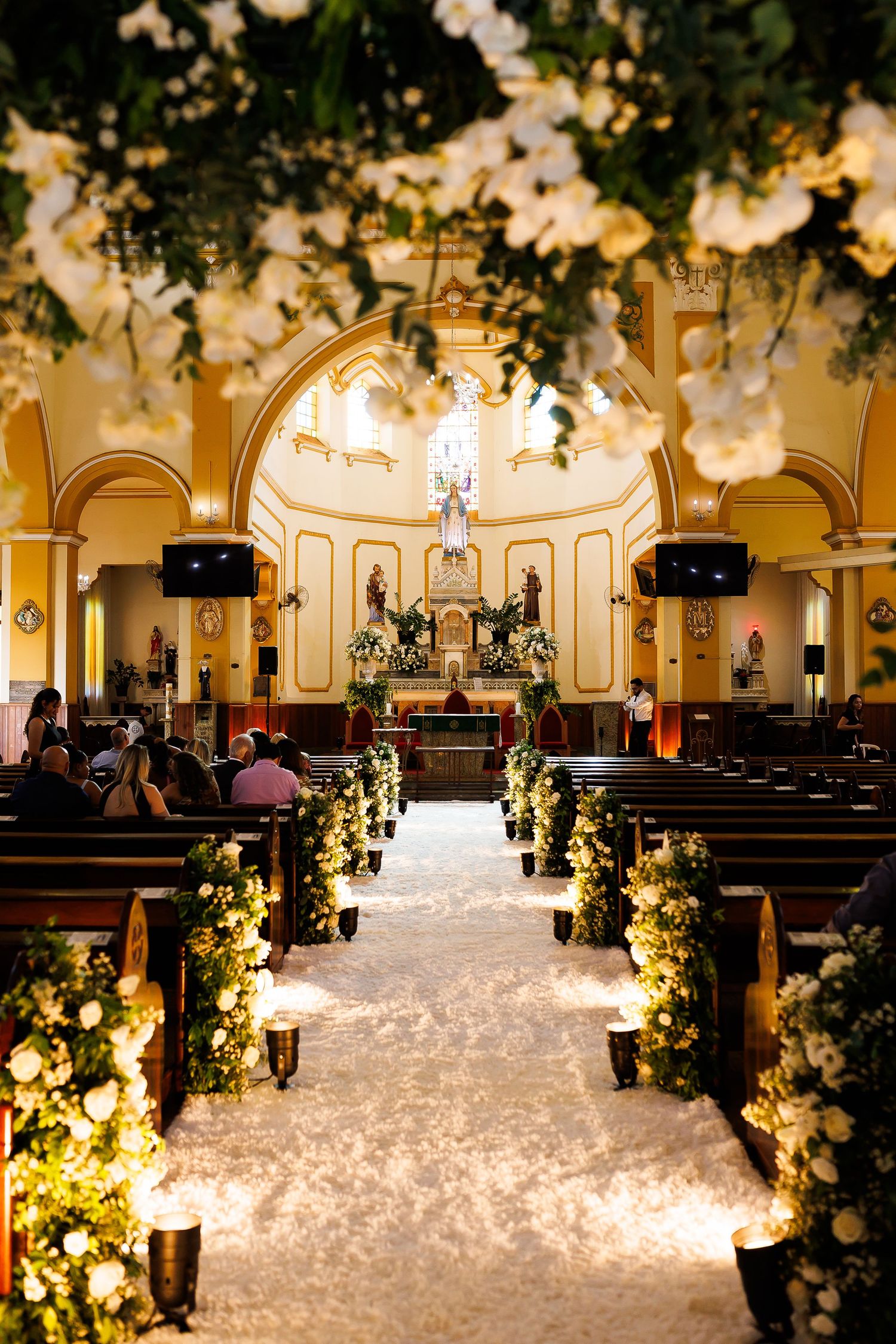 Elegant church wedding aisle adorned with white flowers, greenery, and candlelight leading to a golden altar.