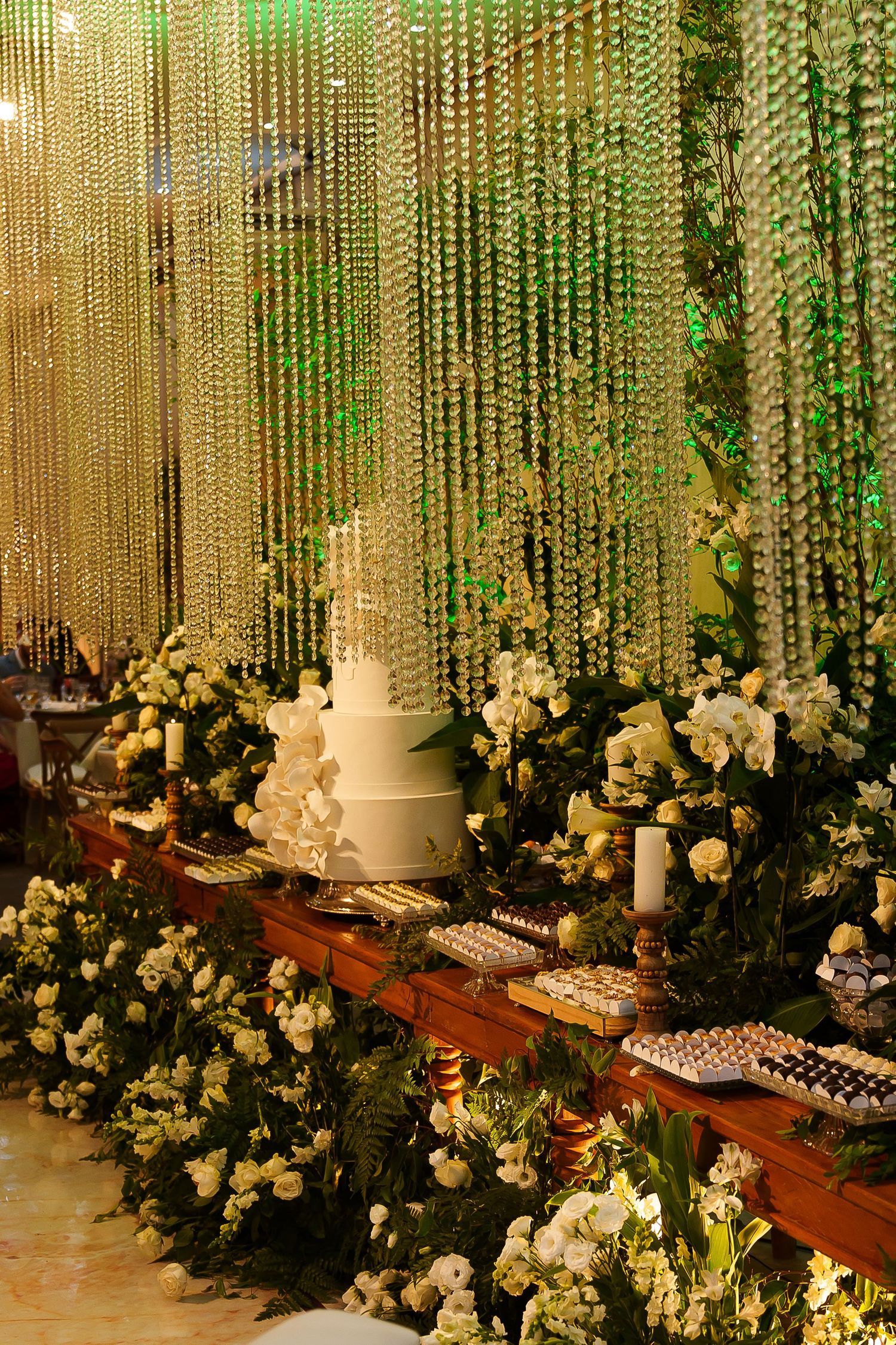 Elegant wedding dessert table with white tiered cake, floral arrangements, and crystal bead curtain backdrop.