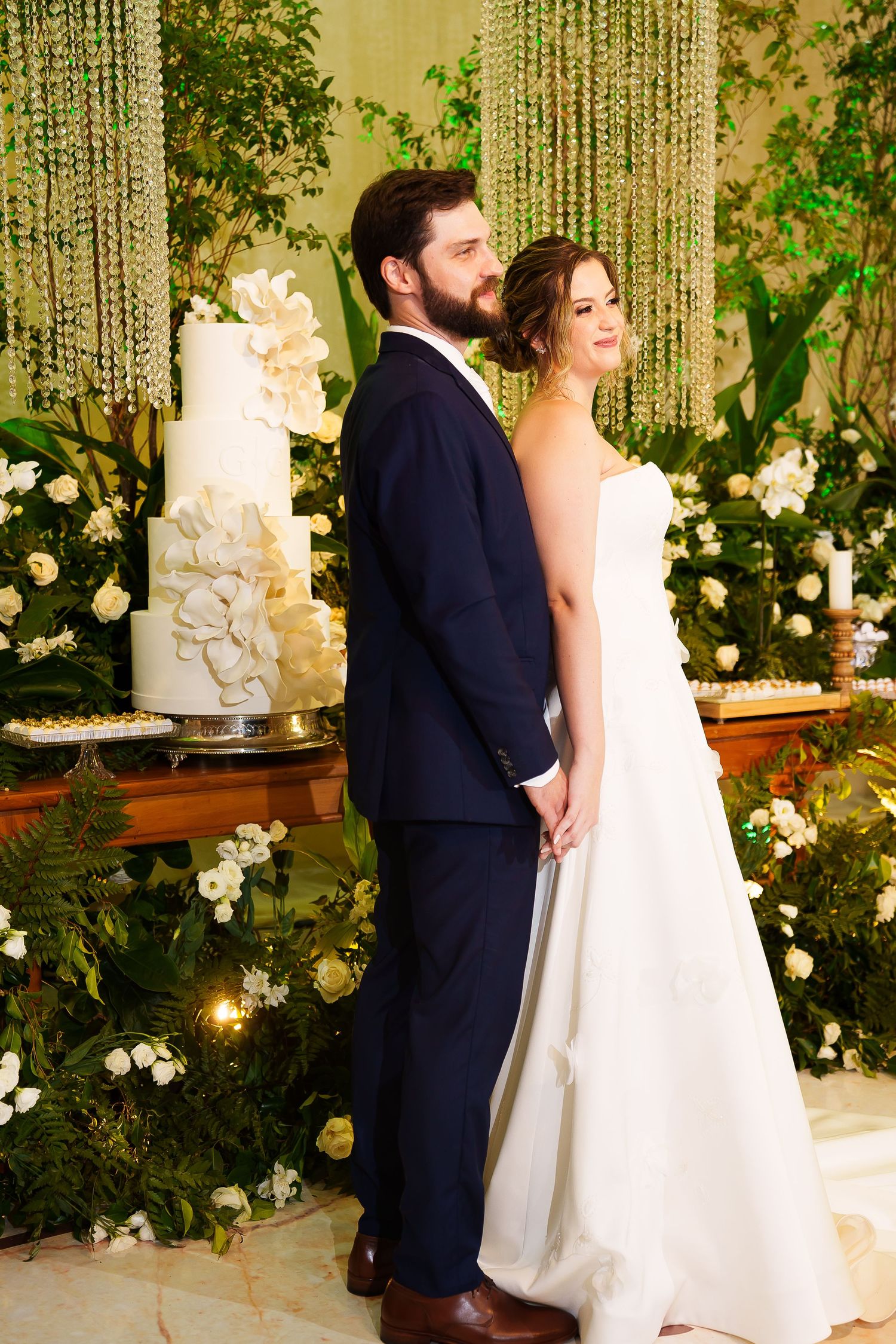 Bride and groom stand back-to-back holding hands near elegant white wedding cake surrounded by lush greenery.