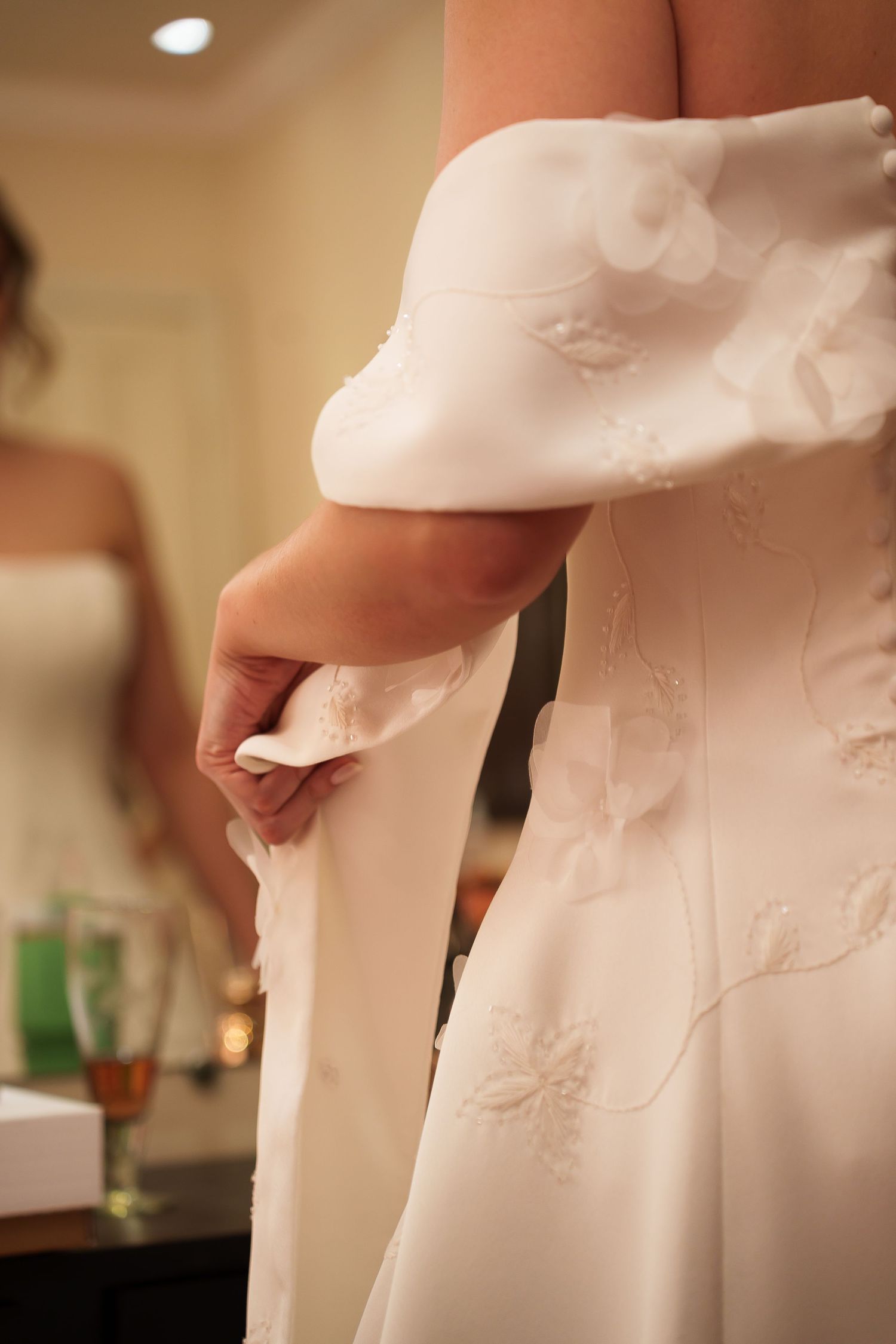 Bride in white embroidered wedding gown adjusting her dress, with mirror reflection visible in background.