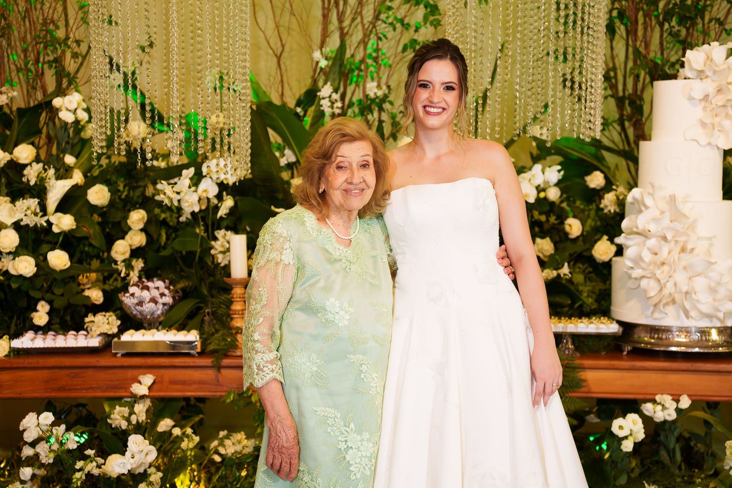 Bride in white gown poses with older woman in green lace dress before floral wedding backdrop with white cake.