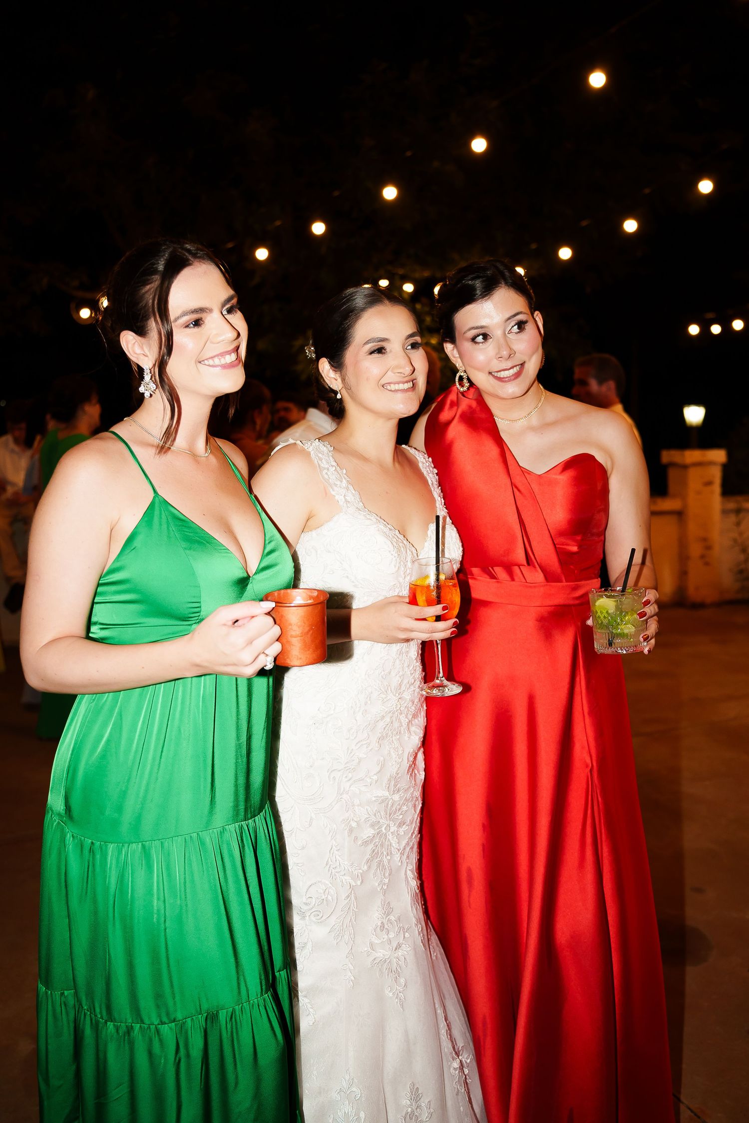 Bride in white lace gown poses with two women in green and red satin dresses at an outdoor evening wedding reception.