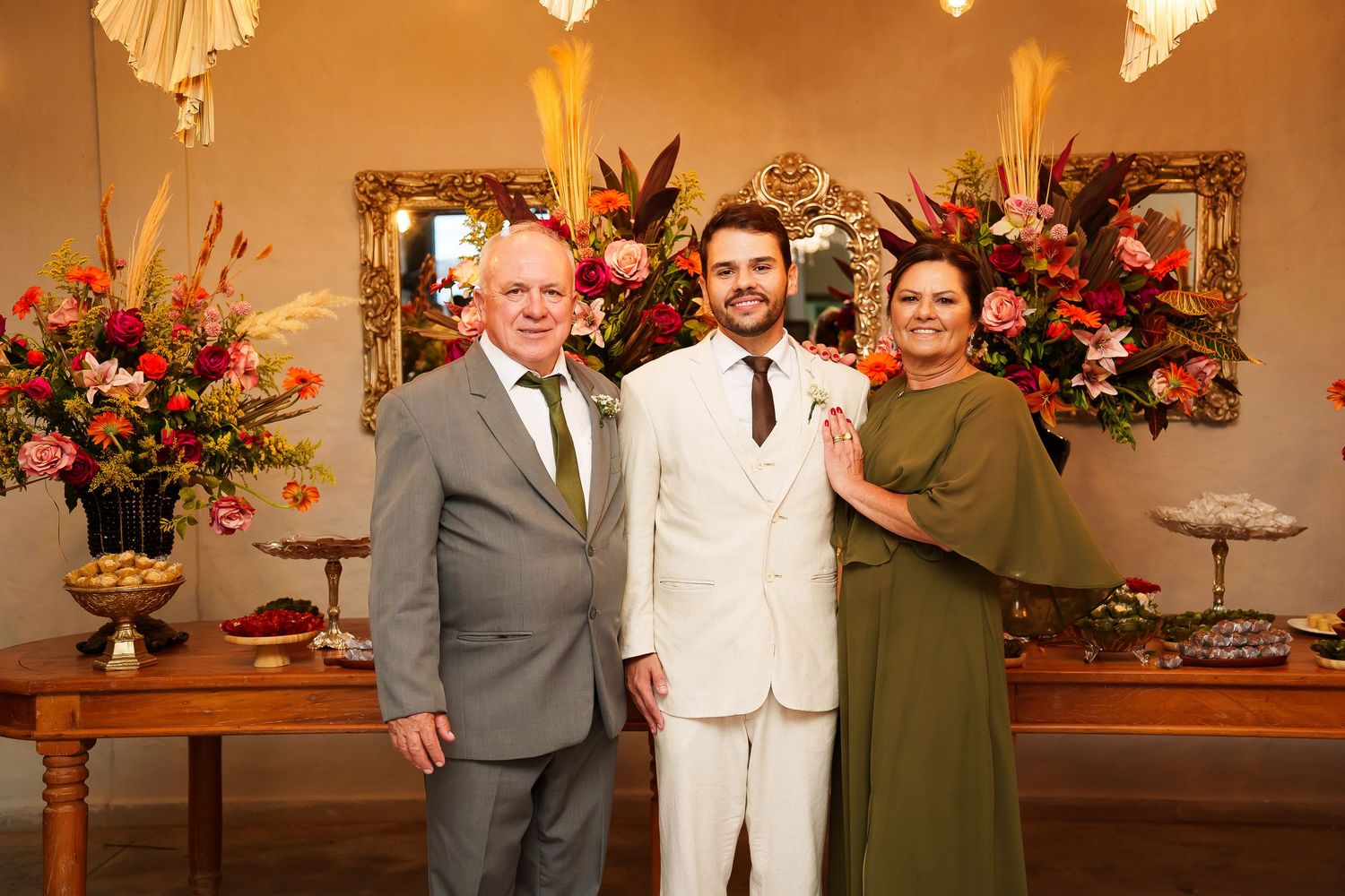Three elegantly dressed people posing together at a wedding reception with colorful floral arrangements in the background.