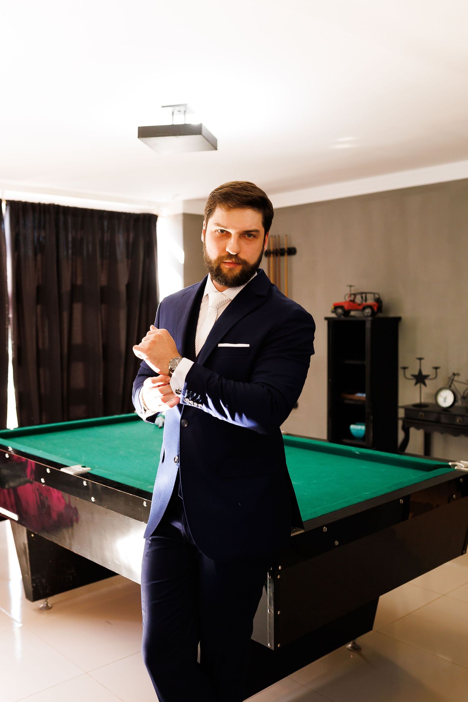 Bearded man in navy blue suit adjusting cufflinks standing beside a green billiard pool table indoors.