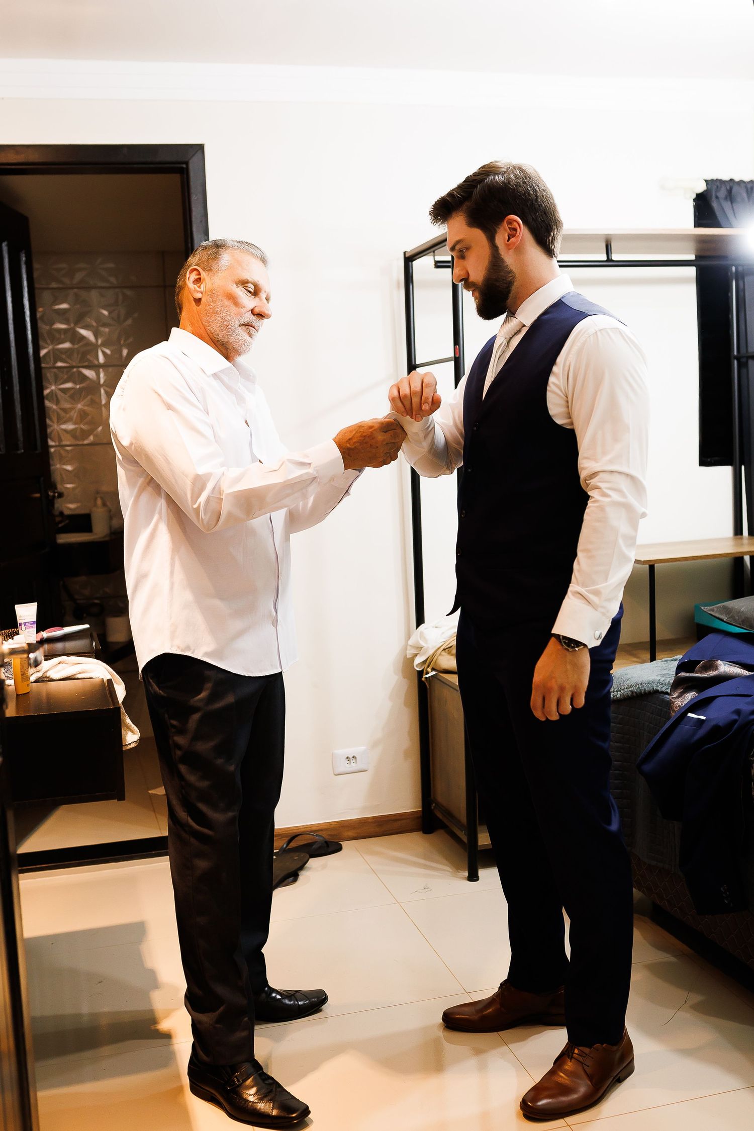 Older man helping young groom adjust his suit vest in a well-lit room before a wedding ceremony.