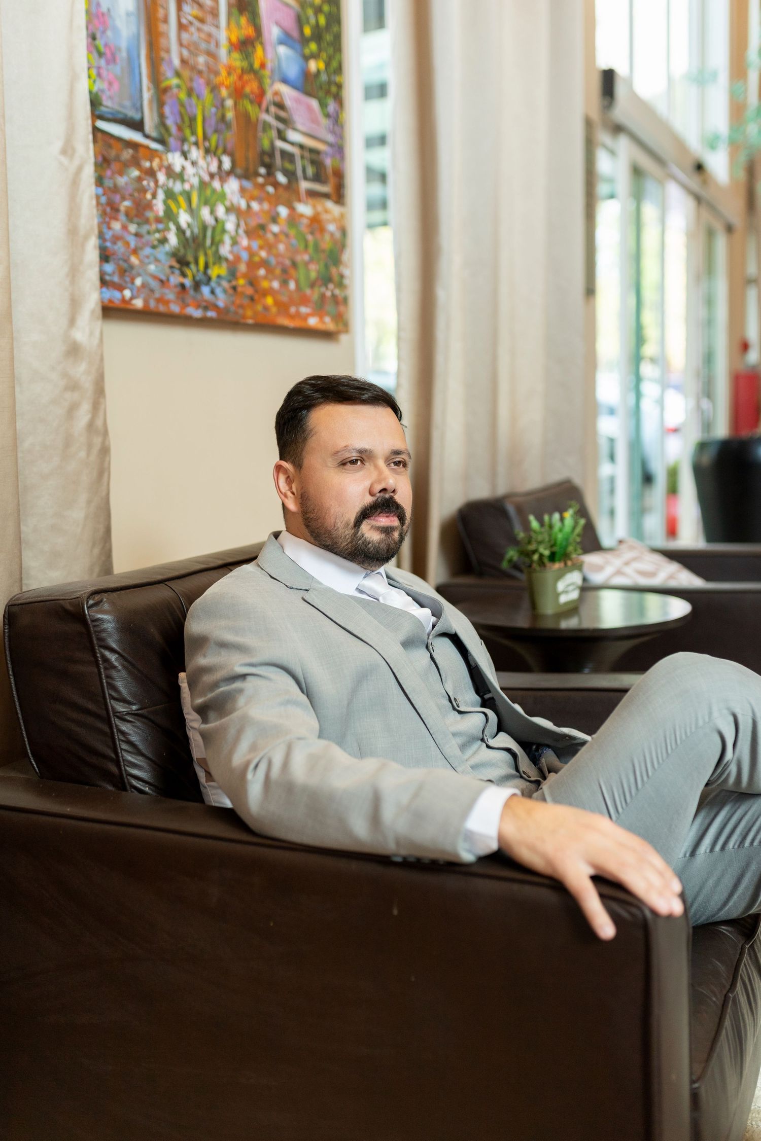 Professional man in gray suit sitting relaxed in leather chair, colorful artwork on wall behind him.