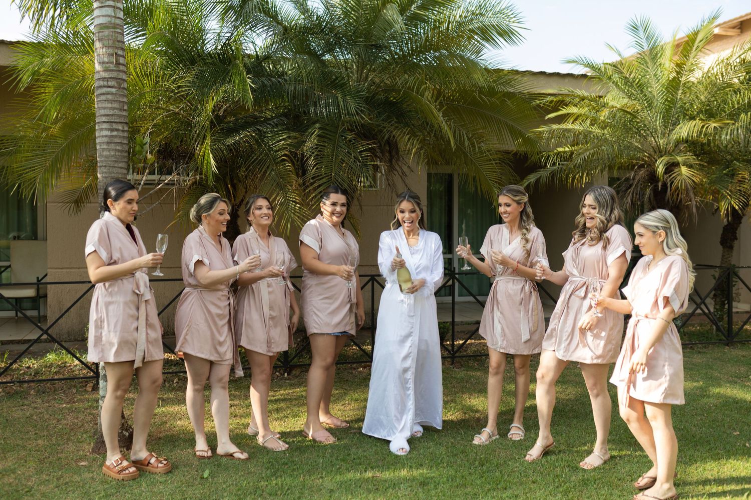 Bride in white robe with bridesmaids in matching pink robes celebrating outdoors near tropical palm trees.