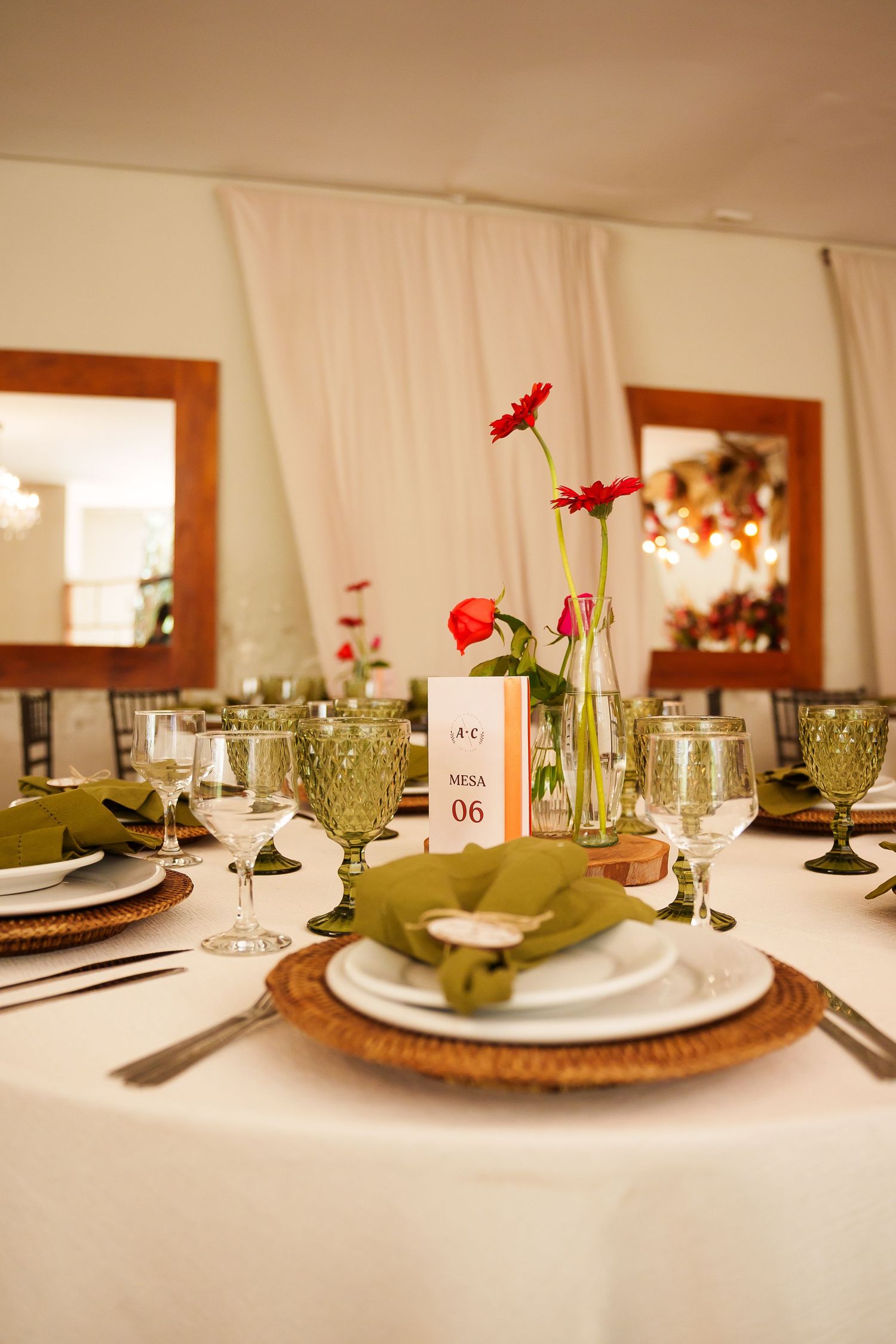 Elegant wedding table setting with green goblets, white plates, red gerbera flowers, and a Mesa 06 table number card.