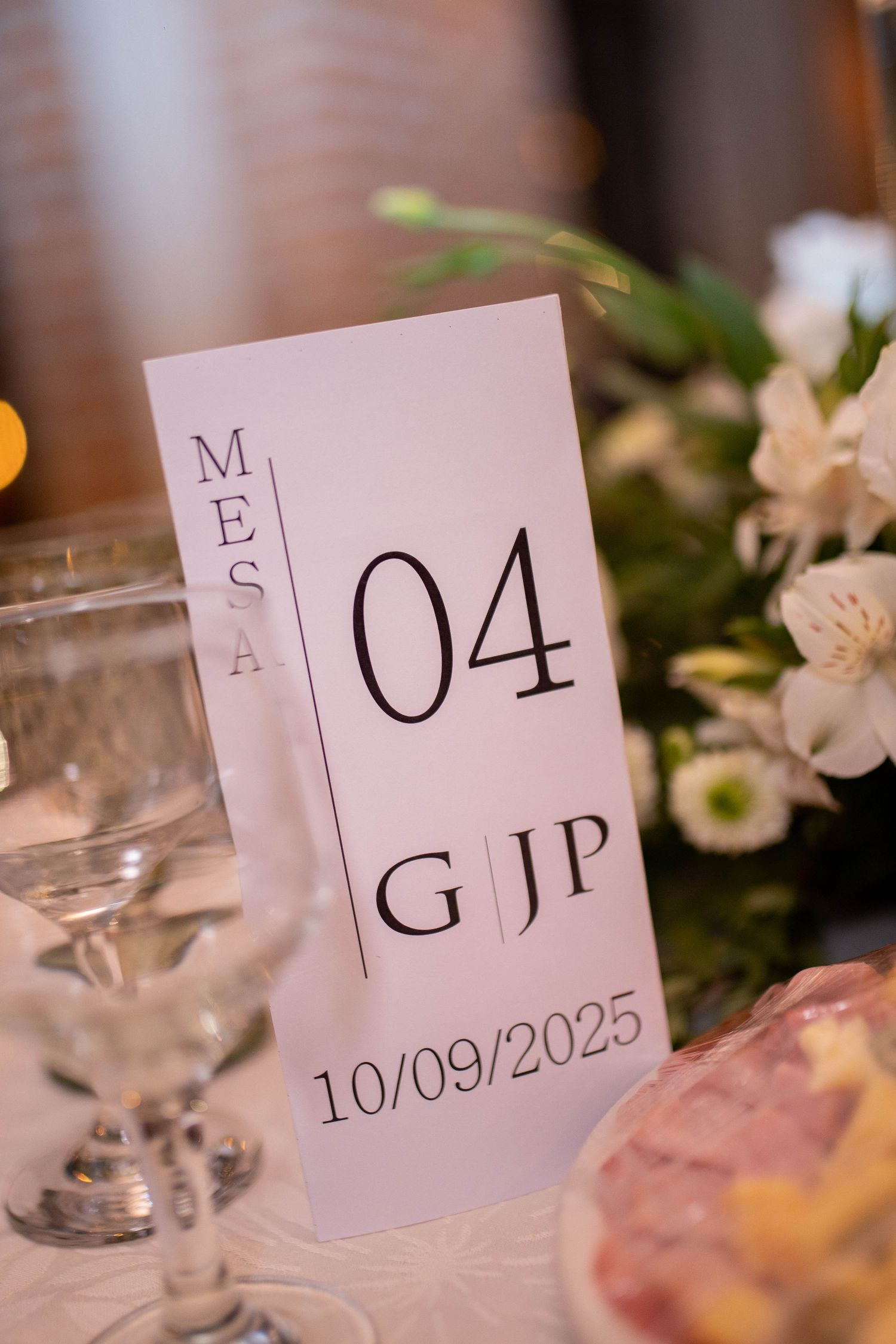 Elegant wedding table number card for Mesa 04 with initials GJP and date 10/09/2025, surrounded by floral decor.