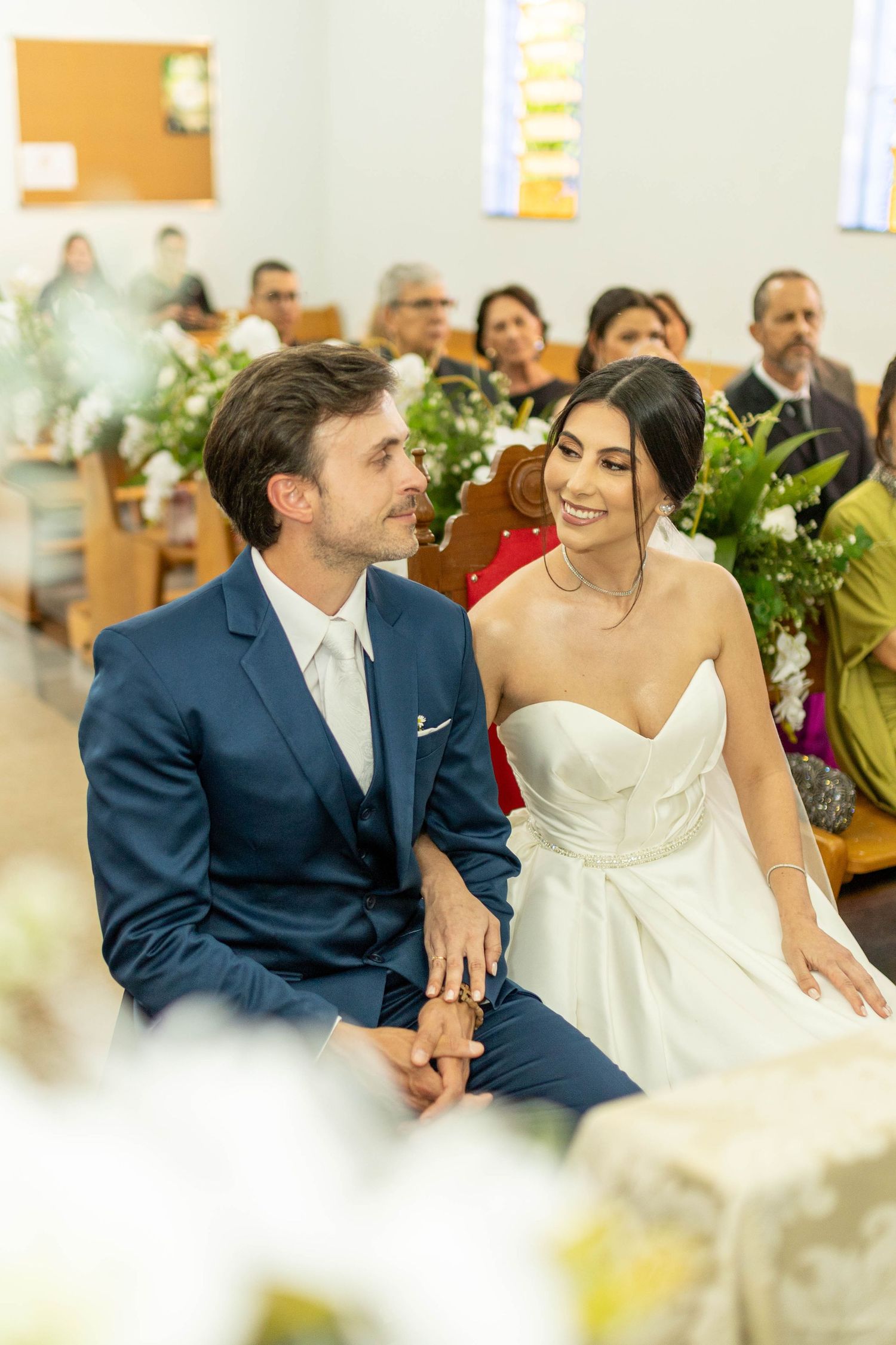 Bride and groom sitting together at church altar during wedding ceremony, smiling lovingly at each other.