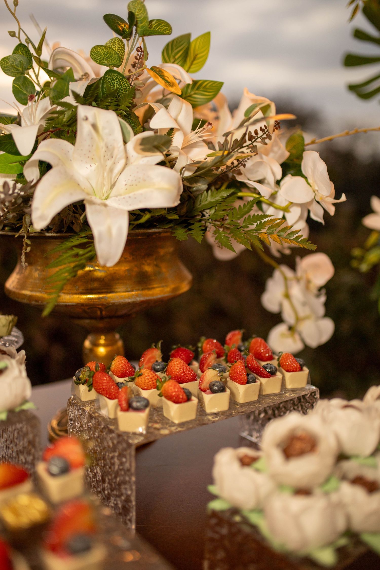 Elegant dessert table with strawberry petit fours, white lily floral arrangement in gold vase, and decorative pastries at an event.