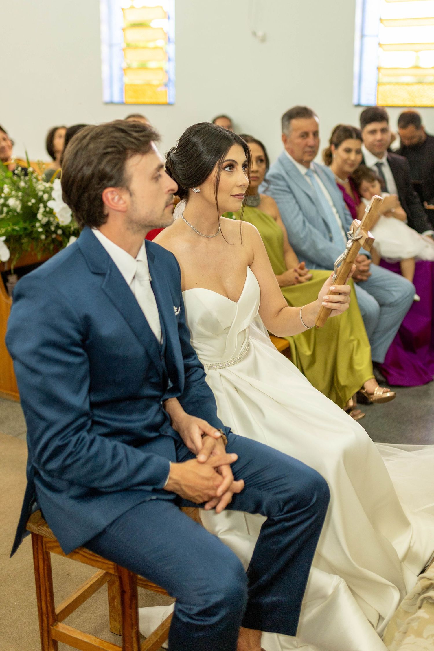 Bride and groom sit together during church wedding ceremony, bride holding candle, guests seated in pews behind them.