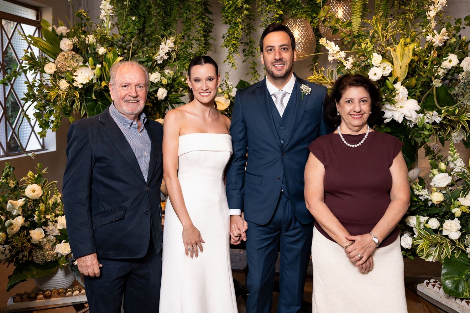 Wedding couple in white dress and suit pose with two guests before floral backdrop of white roses and greenery.